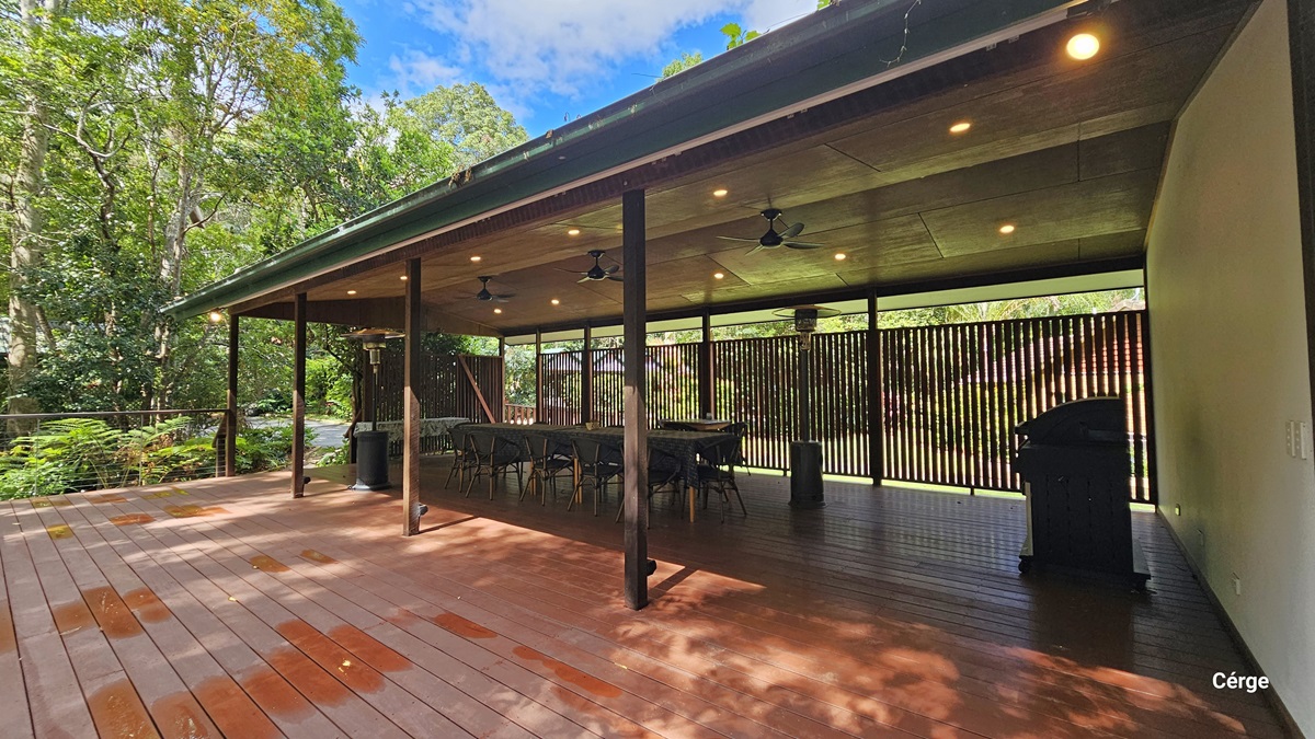 The expansive Rainforest Deck boasts brown timber plank flooring, a gently sloped wooden roof in brown, adorned with three ceiling fans, and a portable heater. Tables and chairs are meticulously arranged along the sides and in the central area. On the right, a wall featuring a wooden plank design extends towards the rear. To the left, there is an open verandah secured with metal railings that provide captivating views of the surrounding rainforest. The expansive Rainforest Deck boasts brown timber plank flooring, a gently sloped wooden roof in brown, adorned with three ceiling fans, and a portable heater. Tables and chairs are meticulously arranged along the sides and in the central area. On the right, a wall featuring a wooden plank design extends towards the rear. To the left, there is an open verandah secured with metal railings that provide captivating views of the surrounding rainforest.
