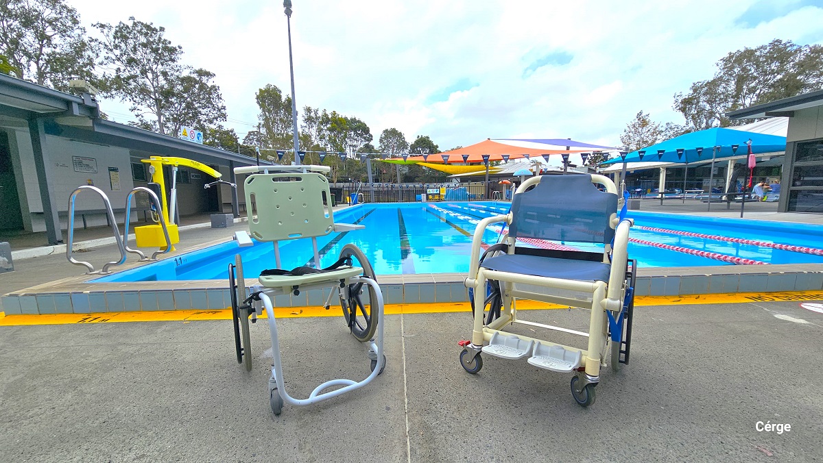 Two aquatic wheelchairs on the pool deck of the 25m, 4-lane outdoor pool. The left aquatic wheelchair has a light green seat and a back support. It has a light blue frame for the foot and a light blue handle for pushing. The right wheelchair has a waterproof cloth cover for seat and back support. It has two-foot support and is composed of cream metal frames. Two aquatic wheelchairs on the pool deck of the 25m, 4-lane outdoor pool. The left aquatic wheelchair has a light green seat and a back support. It has a light blue frame for the foot and a light blue handle for pushing. The right wheelchair has a waterproof cloth cover for seat and back support. It has two-foot support and is composed of cream metal frames.