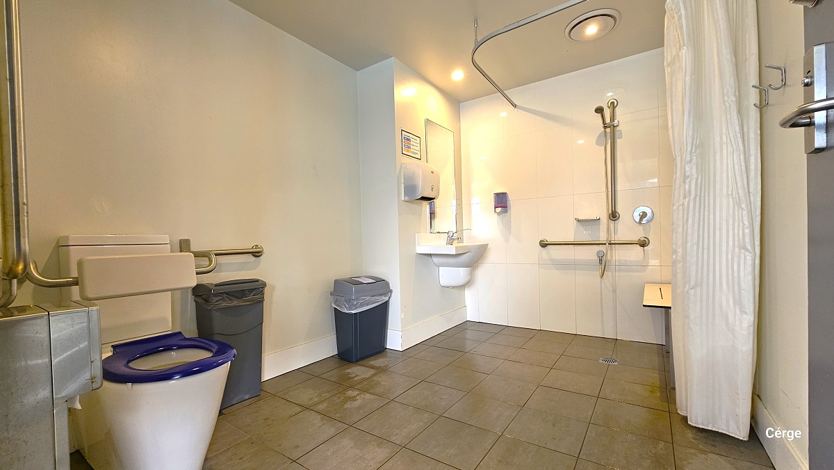 Inside the accessible bathroom at Nerang Aquatic Centre. Near the door is the toilet with a blue toilet cover and white back support. Grab bars are available on the walls near the toilet. On the left of the toilet is the tissue dispenser. There are two grey garbage bins inside the toilet and shower area. The shower has a tall white shower curtain, a wall-mounted shower seat, a shower head with grab bars, and a small basin with a tall wall mirror. The bathroom has a tiled floor with white walls. Inside the accessible bathroom at Nerang Aquatic Centre. Near the door is the toilet with a blue toilet cover and white back support. Grab bars are available on the walls near the toilet. On the left of the toilet is the tissue dispenser. There are two grey garbage bins inside the toilet and shower area. The shower has a tall white shower curtain, a wall-mounted shower seat, a shower head with grab bars, and a small basin with a tall wall mirror. The bathroom has a tiled floor with white walls.