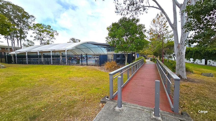 There are two grey rectangular bollards at the start of the red bridge with grey guardrails with strings of wire underneath. There are also red bell-shaped lamp posts next to this bridge. A black fence separates the outdoor amenities from the park. There are two grey rectangular bollards at the start of the red bridge with grey guardrails with strings of wire underneath. There are also red bell-shaped lamp posts next to this bridge. A black fence separates the outdoor amenities from the park.