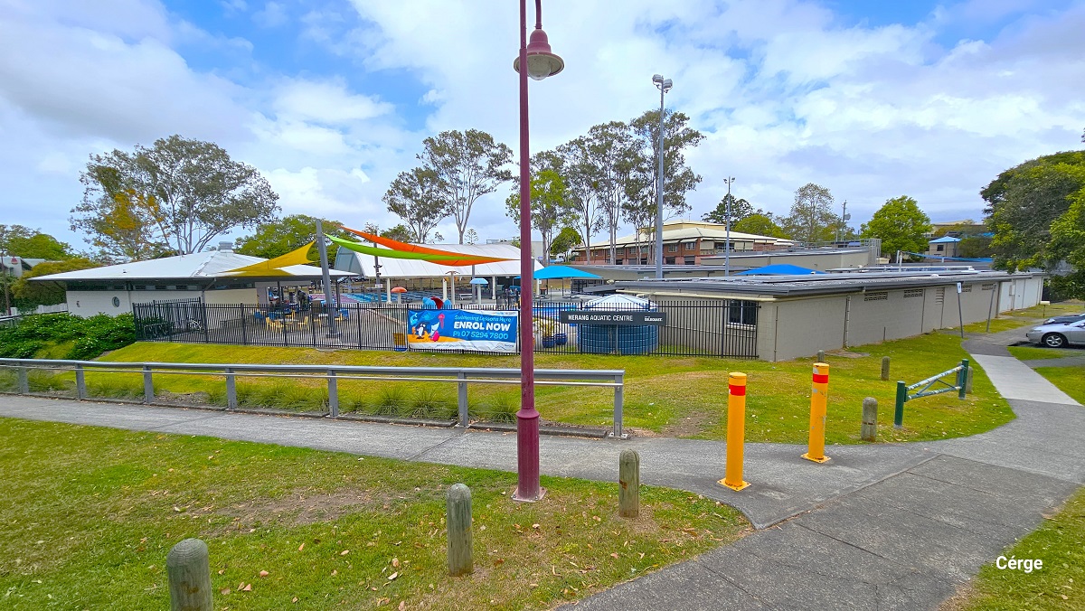 Arrival at Nerang Aquatic Centre from Martin Street with yellow curb marking along an intersection. The sloping road has a middle red brick section and is connected to a middle red and white road markings. Opposite the road is the facility with a small number of designated car spaces marked by white lines, which are occupied by varying brands and colors and cars. Arrival at Nerang Aquatic Centre from Martin Street with yellow curb marking along an intersection. The sloping road has a middle red brick section and is connected to a middle red and white road markings. Opposite the road is the facility with a small number of designated car spaces marked by white lines, which are occupied by varying brands and colors and cars.
