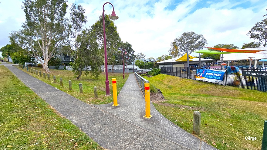 View standing on the start of the concrete pathway with two yellow bollards leading to the entrance and reception desk of Nerang Aquatic Centre. The right side is lined with grey metal handrails with strings of wire underneath, while the left side is red bell-shaped lamp posts. The left section is mostly occupied by grass and tall trees. Hanging on the black perimeter fence is a large blue advertisement and a green signboard detailing the name of the centre. View standing on the start of the concrete pathway with two yellow bollards leading to the entrance and reception desk of Nerang Aquatic Centre. The right side is lined with grey metal handrails with strings of wire underneath, while the left side is red bell-shaped lamp posts. The left section is mostly occupied by grass and tall trees. Hanging on the black perimeter fence is a large blue advertisement and a green signboard detailing the name of the centre.