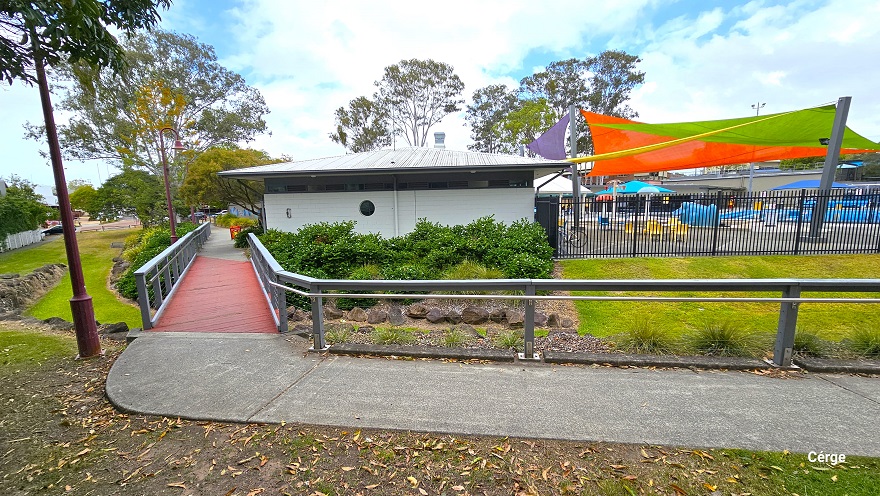 Image shows a red bridge section with grey guardrails connected from the concrete pathway. On both sides is a beautiful landscape of well-maintained grass and a shrub above an arranged rock. Right after the red bridge section is the administration office of a small crème brick building with a prominent hole in the wall. Visible from this position is the colorful sun shades covering the pool. Image shows a red bridge section with grey guardrails connected from the concrete pathway. On both sides is a beautiful landscape of well-maintained grass and a shrub above an arranged rock. Right after the red bridge section is the administration office of a small crème brick building with a prominent hole in the wall. Visible from this position is the colorful sun shades covering the pool.