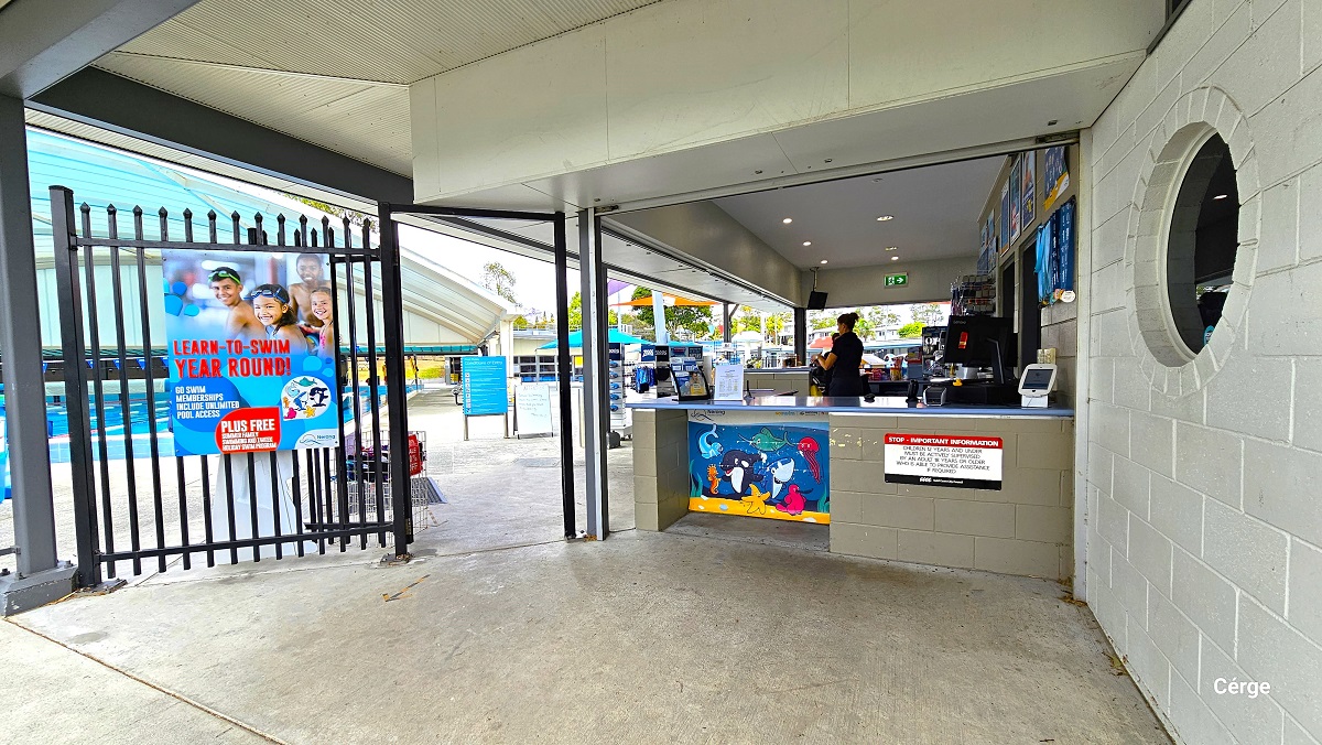A low-height reception counter adjoining a tall black pool fence creates an entry point to pay or scan for admittance to the Nerang Aquatic Centre. There is a large dark blue signboard containing the opening hours, the different facilities, and their rules and conditions and a red bell-shaped lamp post near the black fence. On the right is a small, black, open sign and a garden filled with shrubs and trees with abundant foliage. A low-height reception counter adjoining a tall black pool fence creates an entry point to pay or scan for admittance to the Nerang Aquatic Centre. There is a large dark blue signboard containing the opening hours, the different facilities, and their rules and conditions and a red bell-shaped lamp post near the black fence. On the right is a small, black, open sign and a garden filled with shrubs and trees with abundant foliage.