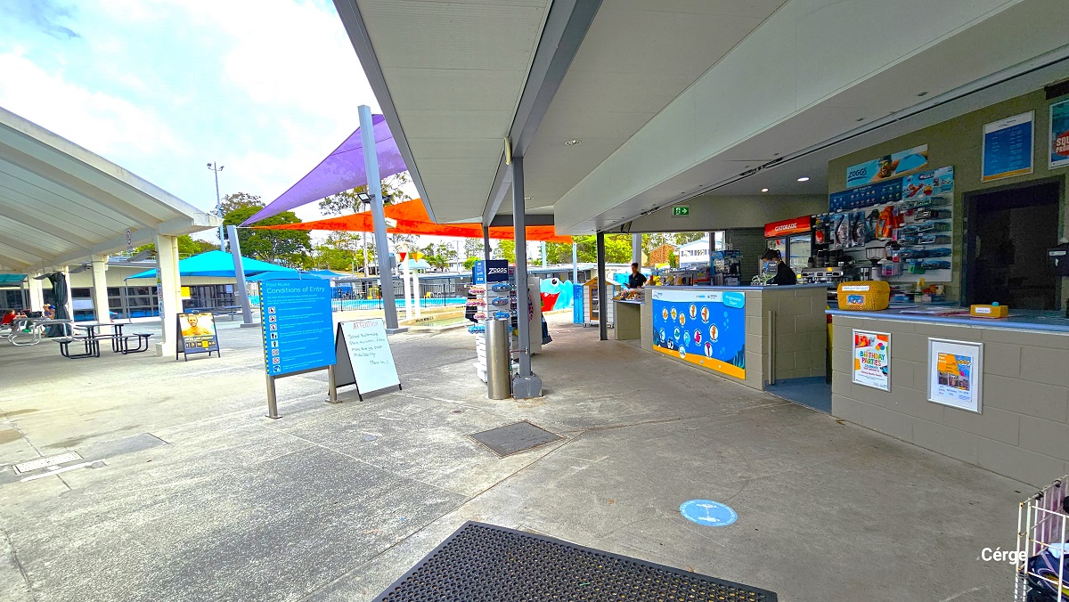 There is a small retail area with aquatic items, snacks, and beverages after entering through the gate and admittance through the reception desk. It has a grey brick reception desk with a light blue countertop designed with colorful animated ocean creatures. Directly in front is the blue sign board for the conditions of entry to the pool and a small whiteboard. From here, guests can see the different outdoor aquatic amenities. There is a small retail area with aquatic items, snacks, and beverages after entering through the gate and admittance through the reception desk. It has a grey brick reception desk with a light blue countertop designed with colorful animated ocean creatures. Directly in front is the blue sign board for the conditions of entry to the pool and a small whiteboard. From here, guests can see the different outdoor aquatic amenities.