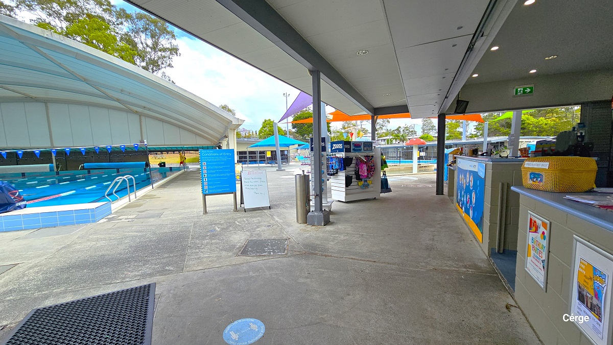 View standing from the retail area in Nerang Aquatic Centre. To the left is the main 10-lane, 25m pool with a white dome structure. The pool has red, blue, and white floating rope lanes, a string of triangular blue flags overhang, and a pool ladder distinguished by the curvy handle. The start of the pool has an elevated blue and white tiled floor and diving blocks on top with a blue cover. Directly ahead is a small splash park and toddler wading pool. View standing from the retail area in Nerang Aquatic Centre. To the left is the main 10-lane, 25m pool with a white dome structure. The pool has red, blue, and white floating rope lanes, a string of triangular blue flags overhang, and a pool ladder distinguished by the curvy handle. The start of the pool has an elevated blue and white tiled floor and diving blocks on top with a blue cover. Directly ahead is a small splash park and toddler wading pool.