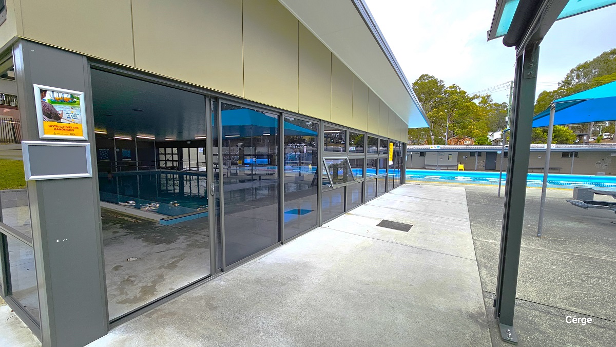 Low-set grey building with tinted glass walls and sheet metal roofing of the indoor programs pool at Nerang Aquatic Centre. It has a cream-colored wall and a white ceiling. On the left grey foundation support are two silver frames with graphics. The indoor pool is behind the large blue tent with a grey picnic table. Low-set grey building with tinted glass walls and sheet metal roofing of the indoor programs pool at Nerang Aquatic Centre. It has a cream-colored wall and a white ceiling. On the left grey foundation support are two silver frames with graphics. The indoor pool is behind the large blue tent with a grey picnic table.