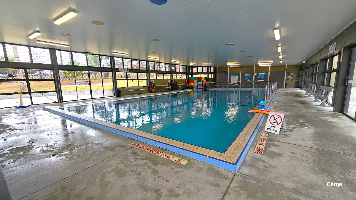 Near the pool edge is a warning sign for shallow water of 0.9 m painted on the floor. There is also a small white sign board for the No Diving warning. On both sides of the pool deck are the benches placed against the wall. On the grey wall to the back, divided by white lines, are adverts. There is a small section on the left wall to the back with colorful cubes. Near the pool edge is a warning sign for shallow water of 0.9 m painted on the floor. There is also a small white sign board for the No Diving warning. On both sides of the pool deck are the benches placed against the wall. On the grey wall to the back, divided by white lines, are adverts. There is a small section on the left wall to the back with colorful cubes.