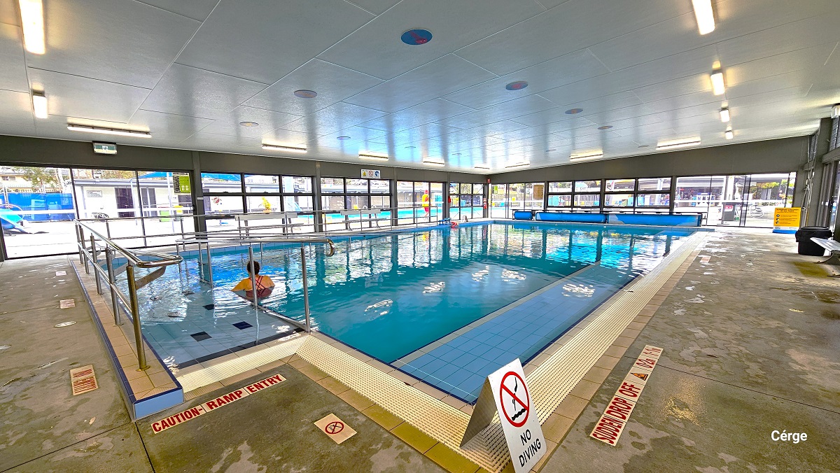 View from the ramp access of the indoor programs pool. At the entry is a red letter warning for “Caution - Ramp Entry.” A no entry sign is placed on the pool deck directly in front of the ramp access with handrails. Near the No Diving white sign board is another caution for “Sudden drop off” detailed on the right pool deck. Further to the back is a black garbage bin. There are blue circles on the ceiling. View from the ramp access of the indoor programs pool. At the entry is a red letter warning for “Caution - Ramp Entry.” A no entry sign is placed on the pool deck directly in front of the ramp access with handrails. Near the No Diving white sign board is another caution for “Sudden drop off” detailed on the right pool deck. Further to the back is a black garbage bin. There are blue circles on the ceiling.