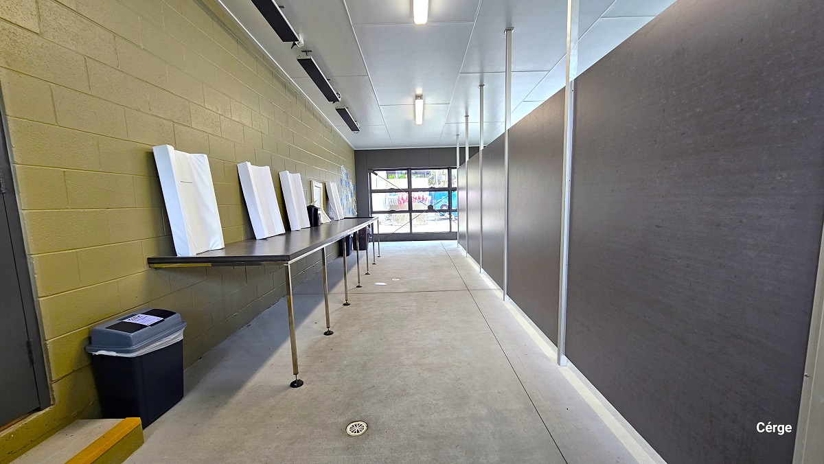 Behind the grey back wall, separated by metal frames connected from the ceiling to the floor. There is a long brown table with white frames on top of it. The table is mounted on the gold brick wall to the left. The cemented floor is smooth, with a white hole for water draining. There is a single-step stair in front of a dark grey door to the left. There are also dark grey rectangular shapes on the ceiling near the edge. Behind the grey back wall, separated by metal frames connected from the ceiling to the floor. There is a long brown table with white frames on top of it. The table is mounted on the gold brick wall to the left. The cemented floor is smooth, with a white hole for water draining. There is a single-step stair in front of a dark grey door to the left. There are also dark grey rectangular shapes on the ceiling near the edge.