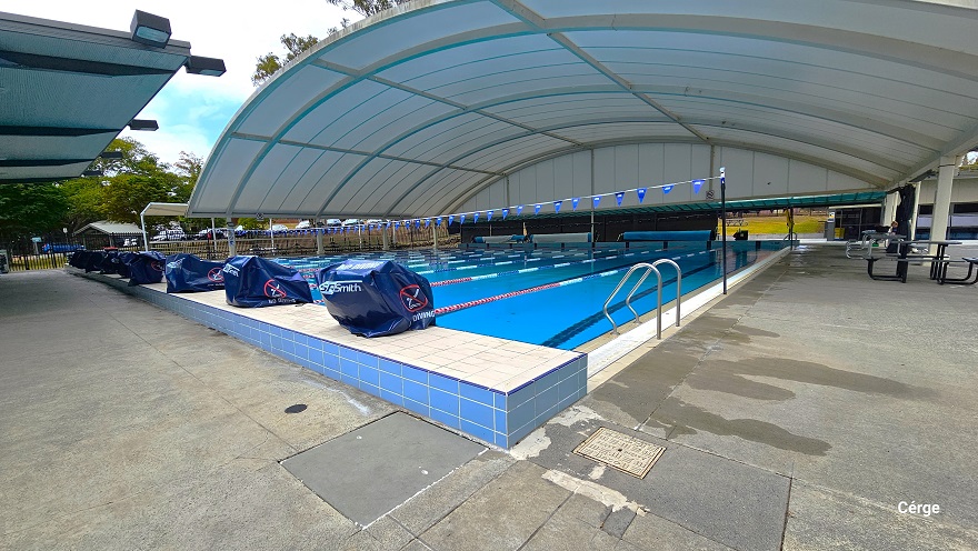 The 25m, 10 lane pool with a large white shaded doom structure at the outdoor aquatic center in Nerang Aquatic. There are two strings of triangular blue flags wrapped around the black poles on the opposite side of the pool. Diving blocks with safety cover are placed on each lane, separated by red, blue, and white floating rope lanes. It has a wide pool deck with three long rolls of blue waterproof tarpaulin at the back. The 25m, 10 lane pool with a large white shaded doom structure at the outdoor aquatic center in Nerang Aquatic. There are two strings of triangular blue flags wrapped around the black poles on the opposite side of the pool. Diving blocks with safety cover are placed on each lane, separated by red, blue, and white floating rope lanes. It has a wide pool deck with three long rolls of blue waterproof tarpaulin at the back.