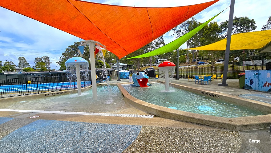 There are red, light green, and yellow shade sails protecting the splash park, toddler wading pool, and the seating area. There is a red mushroom fountain, a large blue frog with a short red water slide, a light blue mushroom fountain, and a cone bucket area with red, blue, and yellow colors. The seating section to the far right has yellow and light blue plastic chairs and tables. There are red, light green, and yellow shade sails protecting the splash park, toddler wading pool, and the seating area. There is a red mushroom fountain, a large blue frog with a short red water slide, a light blue mushroom fountain, and a cone bucket area with red, blue, and yellow colors. The seating section to the far right has yellow and light blue plastic chairs and tables.