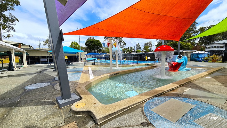 The image shows the Nerang Aquatic Centre's outdoor pool area, featuring a 25m, 10-lane pool covered by a white dome on the left side. There are red and light green shade sails protecting the splash park and toddler wading pool. Water features include a red mushroom fountain, a large blue frog water slide, a light blue mushroom fountain, and a cone bucket area with red, blue, and yellow colors. At the back, there are two large blue tents and a 25m, 4-lane pool separated by a black fence. The image shows the Nerang Aquatic Centre's outdoor pool area, featuring a 25m, 10-lane pool covered by a white dome on the left side. There are red and light green shade sails protecting the splash park and toddler wading pool. Water features include a red mushroom fountain, a large blue frog water slide, a light blue mushroom fountain, and a cone bucket area with red, blue, and yellow colors. At the back, there are two large blue tents and a 25m, 4-lane pool separated by a black fence.