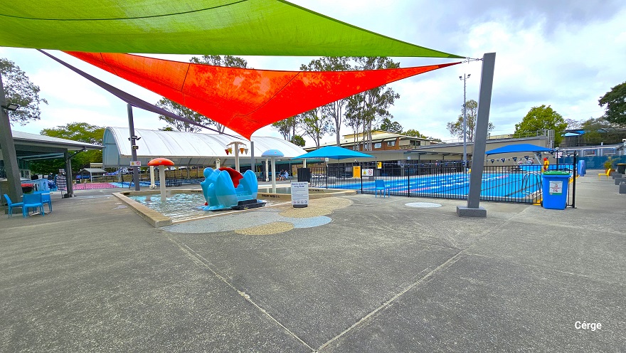 View near the seating section next to the Splash park and Toddler wading pool. The red, light green and purple shade sails are wrapped around the large metal rectangular poles. Splash Park and the Toddler wading pool are separated from the 25m, 4-lane pool by a low black fence. It also has a blue triangular flag, floating rope lanes, and pool ladders. Around the pool deck are yellow plastic chairs. View near the seating section next to the Splash park and Toddler wading pool. The red, light green and purple shade sails are wrapped around the large metal rectangular poles. Splash Park and the Toddler wading pool are separated from the 25m, 4-lane pool by a low black fence. It also has a blue triangular flag, floating rope lanes, and pool ladders. Around the pool deck are yellow plastic chairs.