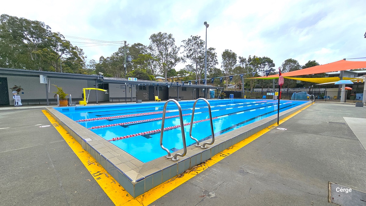 View on the right side of the 25m, 4-lane pool. There is a low-set grey and cream building at the back with long benches placed against the white wall. It has small glass windows for air circulation and illumination. A large, square, white clock can be seen hanging on the roof of the building. The Cérge app boy icon holding a QR code for people with disabilities can be found in front of a grey door near the pool. View on the right side of the 25m, 4-lane pool. There is a low-set grey and cream building at the back with long benches placed against the white wall. It has small glass windows for air circulation and illumination. A large, square, white clock can be seen hanging on the roof of the building. The Cérge app boy icon holding a QR code for people with disabilities can be found in front of a grey door near the pool.