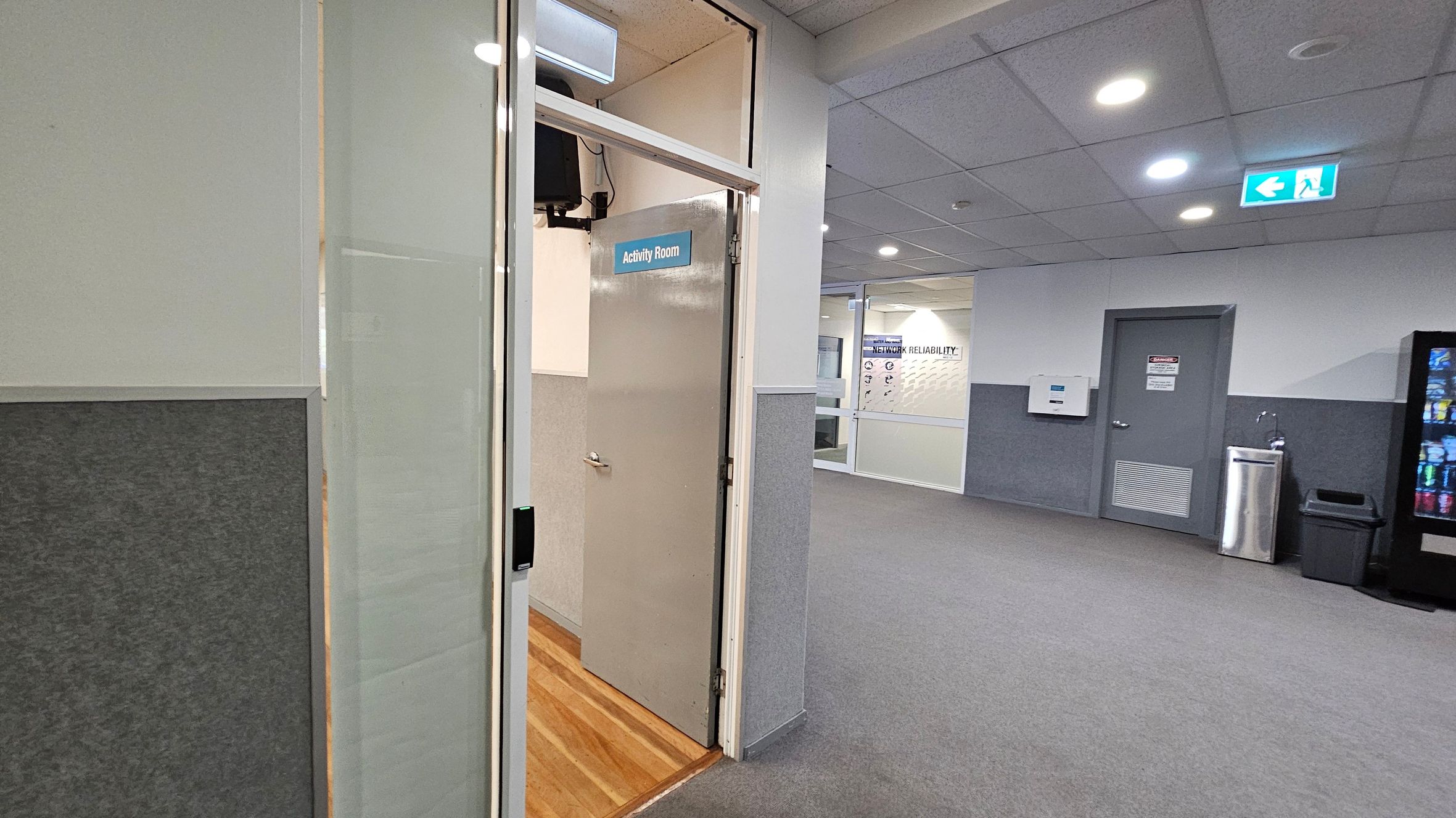 A hallway with grey carpet and a ceiling with recessed lights. On the left, an open door labelled "Activity Room" with a wooden floor inside. On the right, a vending machine, a water fountain, and a waste bin are visible. A door with a warning sign is next to these items. In the background, a glass wall with a poster about network reliability is visible. An emergency exit sign is on the ceiling. A hallway with grey carpet and a ceiling with recessed lights. On the left, an open door labelled "Activity Room" with a wooden floor inside. On the right, a vending machine, a water fountain, and a waste bin are visible. A door with a warning sign is next to these items. In the background, a glass wall with a poster about network reliability is visible. An emergency exit sign is on the ceiling.