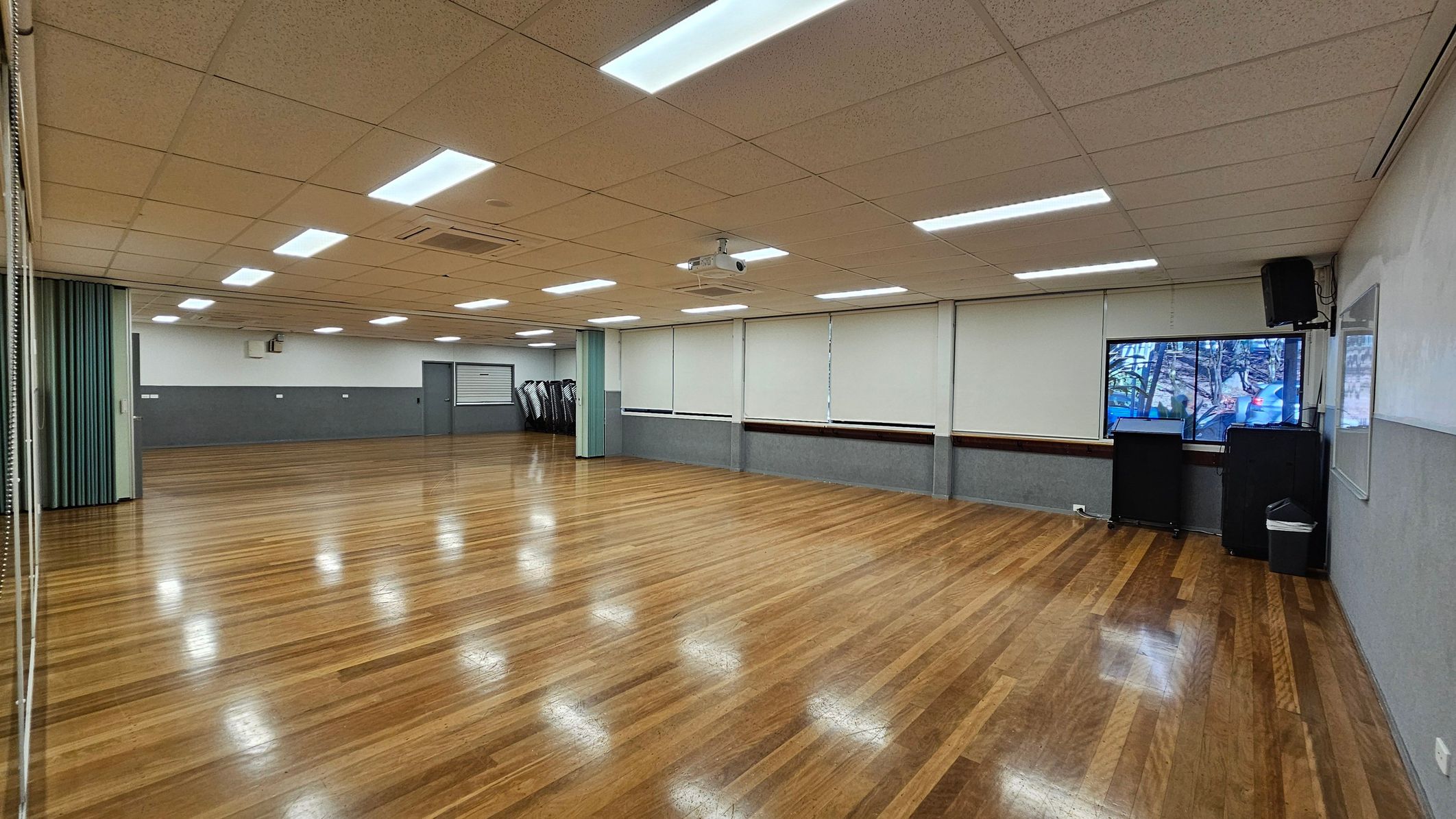 A spacious room with polished wooden floors and a suspended ceiling with fluorescent lights. On the right, there is a window with blinds, a black speaker, and a whiteboard. In the background, a stack of chairs is visible near a closed door. The left side features a folding partition. A projector is mounted on the ceiling. A spacious room with polished wooden floors and a suspended ceiling with fluorescent lights. On the right, there is a window with blinds, a black speaker, and a whiteboard. In the background, a stack of chairs is visible near a closed door. The left side features a folding partition. A projector is mounted on the ceiling.