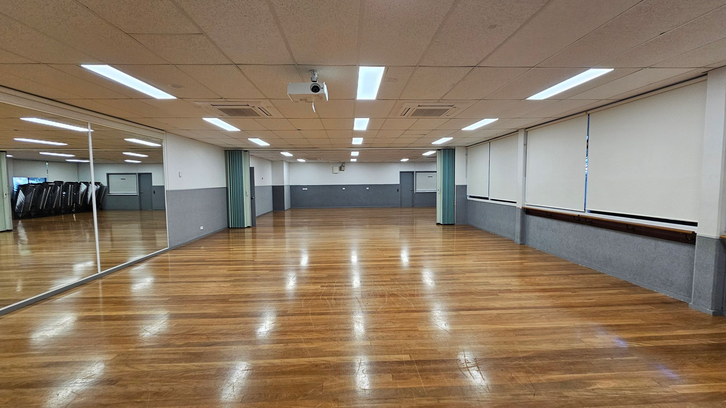 A spacious room with a polished wooden floor and a ceiling with fluorescent lights. On the left, a large mirror reflects the room, showing stacked chairs in the corner. The right side features windows covered with white blinds. The back wall has a grey lower half and white upper half, with a projector mounted on the ceiling. The room appears to be a multipurpose space, possibly for dance or exercise classes. A spacious room with a polished wooden floor and a ceiling with fluorescent lights. On the left, a large mirror reflects the room, showing stacked chairs in the corner. The right side features windows covered with white blinds. The back wall has a grey lower half and white upper half, with a projector mounted on the ceiling. The room appears to be a multipurpose space, possibly for dance or exercise classes.