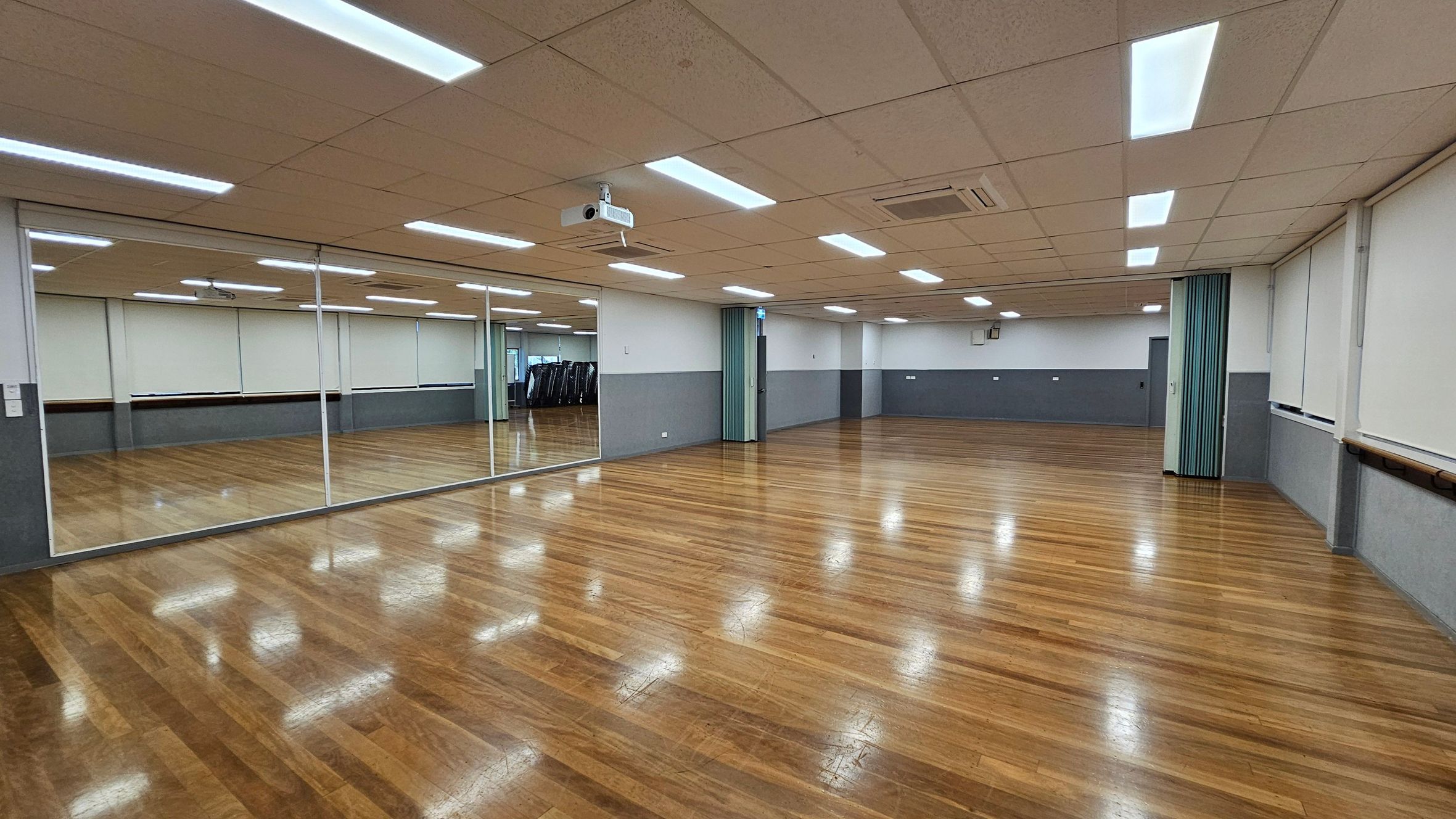 A spacious room with a polished wooden floor and a ceiling with recessed lighting. On the left, a large wall of mirrors reflects the room, enhancing the sense of space. The right side features a series of windows with blinds and a ballet barre running along the wall. In the centre, a projector is mounted on the ceiling. The walls are painted in a two-tone scheme, with a lighter colour on top and a darker shade on the bottom. A spacious room with a polished wooden floor and a ceiling with recessed lighting. On the left, a large wall of mirrors reflects the room, enhancing the sense of space. The right side features a series of windows with blinds and a ballet barre running along the wall. In the centre, a projector is mounted on the ceiling. The walls are painted in a two-tone scheme, with a lighter colour on top and a darker shade on the bottom.