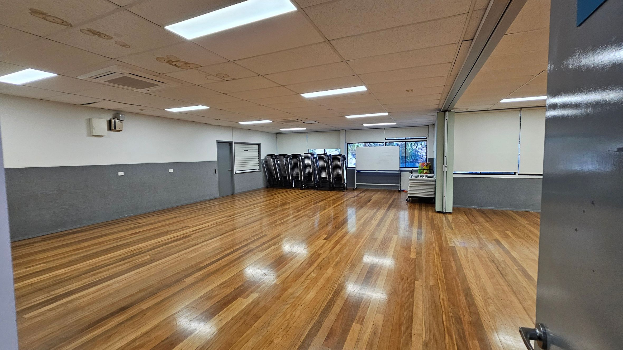 A spacious room with polished wooden floors and a white ceiling with fluorescent lights. On the left, a grey and white wall with a closed door and a small window. In the centre, a stack of chairs and a whiteboard near large windows with blinds. On the right, a trolley with items on top. The room is well-lit and appears to be a multipurpose space. A spacious room with polished wooden floors and a white ceiling with fluorescent lights. On the left, a grey and white wall with a closed door and a small window. In the centre, a stack of chairs and a whiteboard near large windows with blinds. On the right, a trolley with items on top. The room is well-lit and appears to be a multipurpose space.