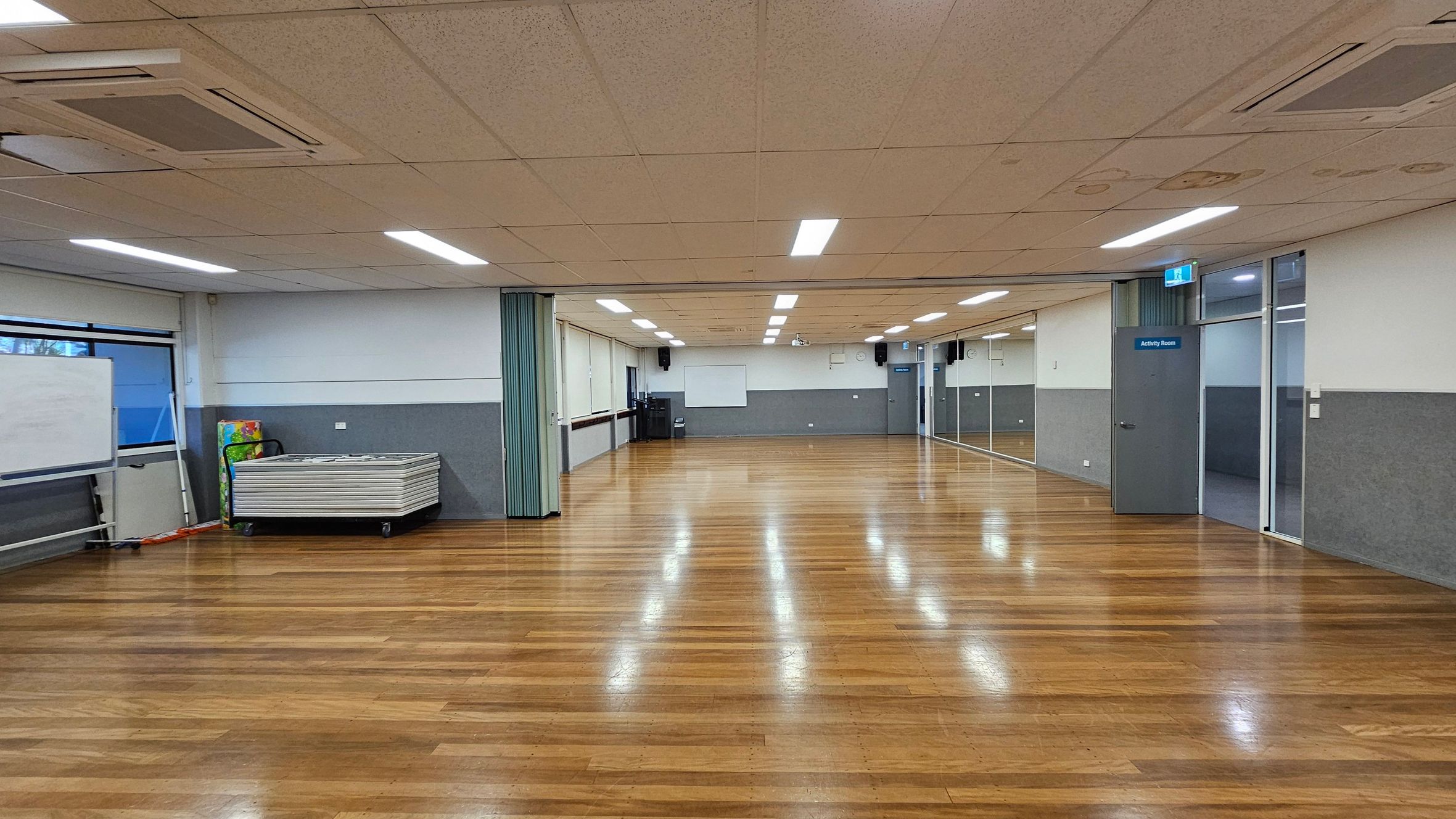A spacious room with wooden flooring and a suspended ceiling with fluorescent lights. On the left, there's a stack of tables and a whiteboard near a window. The right side features a door labelled "Activity Room" and a wall with large mirrors. The back wall has a whiteboard and a clock. The room is well-lit and appears to be used for activities or meetings. A spacious room with wooden flooring and a suspended ceiling with fluorescent lights. On the left, there's a stack of tables and a whiteboard near a window. The right side features a door labelled "Activity Room" and a wall with large mirrors. The back wall has a whiteboard and a clock. The room is well-lit and appears to be used for activities or meetings.