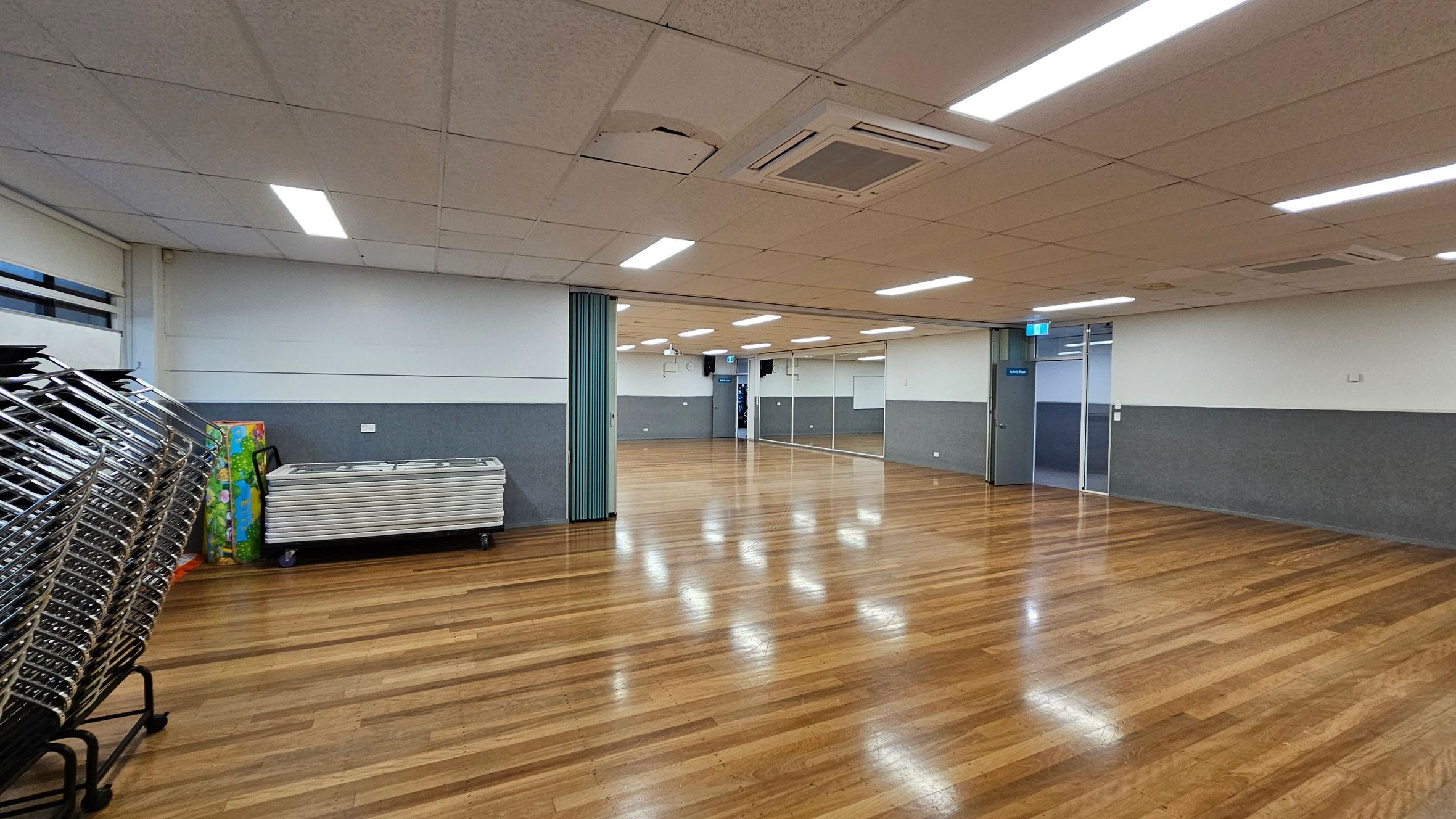 A spacious room with a polished wooden floor and a white ceiling with fluorescent lights. On the left, there are stacks of folded chairs and tables against the wall. The walls are half-painted in grey and white. In the background, there are mirrors on the right wall and a glass door with a blue sign. The room is well-lit and appears to be a multi-purpose space. A spacious room with a polished wooden floor and a white ceiling with fluorescent lights. On the left, there are stacks of folded chairs and tables against the wall. The walls are half-painted in grey and white. In the background, there are mirrors on the right wall and a glass door with a blue sign. The room is well-lit and appears to be a multi-purpose space.