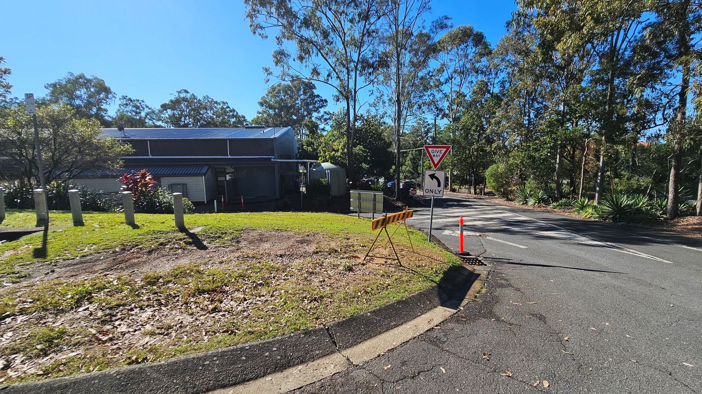 A road curves to the right with a "Give Way" sign and a directional arrow indicating a left turn only. On the left, a building with a metal roof is partially visible, surrounded by trees and shrubs. Concrete bollards line the grassy area in front of the building. A temporary barrier and an orange traffic cone are placed near the road. Tall trees with sparse foliage are in the background, and the road surface is slightly cracked with scattered leaves. A road curves to the right with a "Give Way" sign and a directional arrow indicating a left turn only. On the left, a building with a metal roof is partially visible, surrounded by trees and shrubs. Concrete bollards line the grassy area in front of the building. A temporary barrier and an orange traffic cone are placed near the road. Tall trees with sparse foliage are in the background, and the road surface is slightly cracked with scattered leaves.