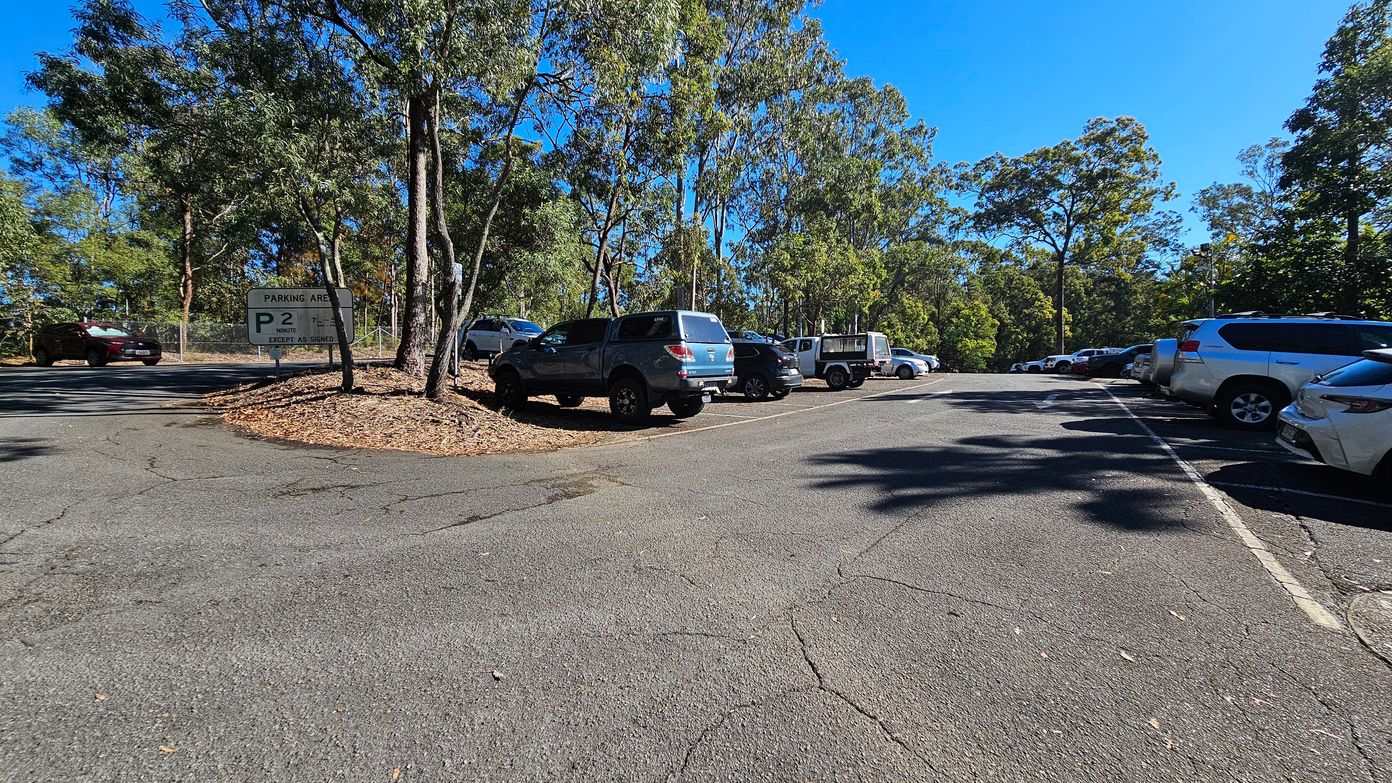 A car park surrounded by tall trees. On the left, a sign indicates a 2-minute parking area. Several vehicles, including a blue SUV and a white utility vehicle, are parked on the left side. On the right, more cars are lined up along the edge of the asphalt surface. The ground is cracked and uneven, with scattered leaves. The background is filled with dense greenery, and the sky is clear and blue. A car park surrounded by tall trees. On the left, a sign indicates a 2-minute parking area. Several vehicles, including a blue SUV and a white utility vehicle, are parked on the left side. On the right, more cars are lined up along the edge of the asphalt surface. The ground is cracked and uneven, with scattered leaves. The background is filled with dense greenery, and the sky is clear and blue.
