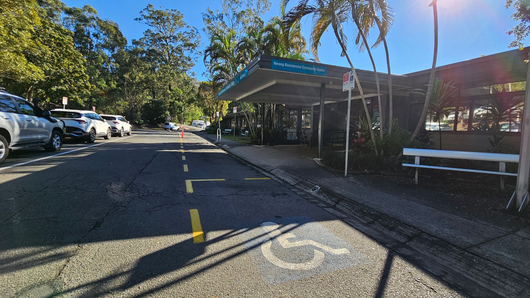 A street scene with a row of parked cars on the left side, including several white vehicles. On the right, a building with a sign reading "Nerang Bicentennial Community Centre" is partially visible, with a covered entrance and a bench nearby. Palm trees and other greenery surround the area. The road has a marked disabled parking space in the foreground, and yellow lines are painted on the asphalt. The background features more trees and a clear blue sky. A street scene with a row of parked cars on the left side, including several white vehicles. On the right, a building with a sign reading "Nerang Bicentennial Community Centre" is partially visible, with a covered entrance and a bench nearby. Palm trees and other greenery surround the area. The road has a marked disabled parking space in the foreground, and yellow lines are painted on the asphalt. The background features more trees and a clear blue sky.