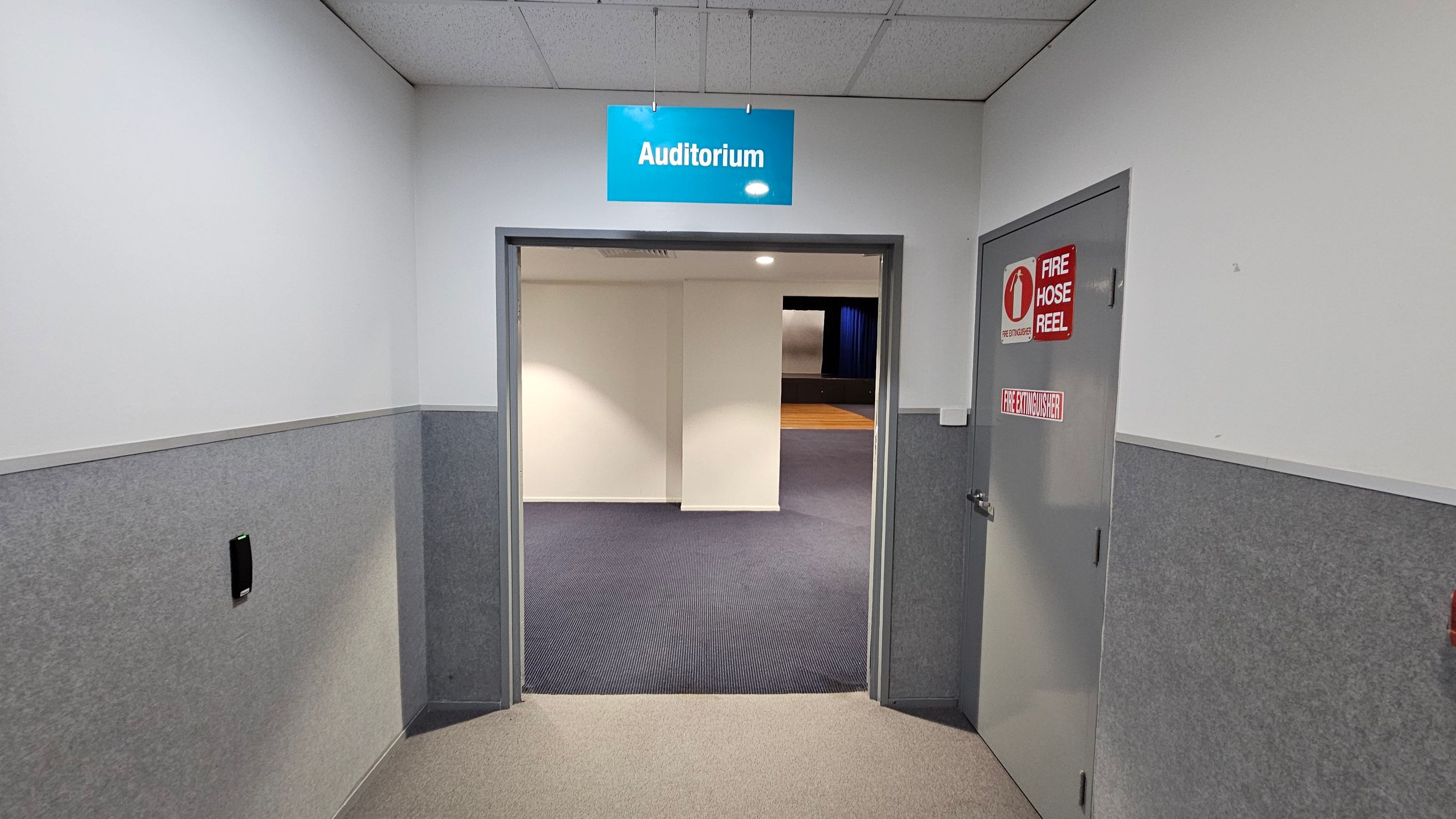 A hallway with grey and white walls leads to an open doorway. Above the doorway, a blue sign reads "Auditorium". To the right, a grey door features red signs for a fire hose reel and fire extinguisher. The floor is carpeted in beige, transitioning to dark grey inside the auditorium. The ceiling is composed of white tiles. In the background, a stage with blue curtains is partially visible. A hallway with grey and white walls leads to an open doorway. Above the doorway, a blue sign reads "Auditorium". To the right, a grey door features red signs for a fire hose reel and fire extinguisher. The floor is carpeted in beige, transitioning to dark grey inside the auditorium. The ceiling is composed of white tiles. In the background, a stage with blue curtains is partially visible.