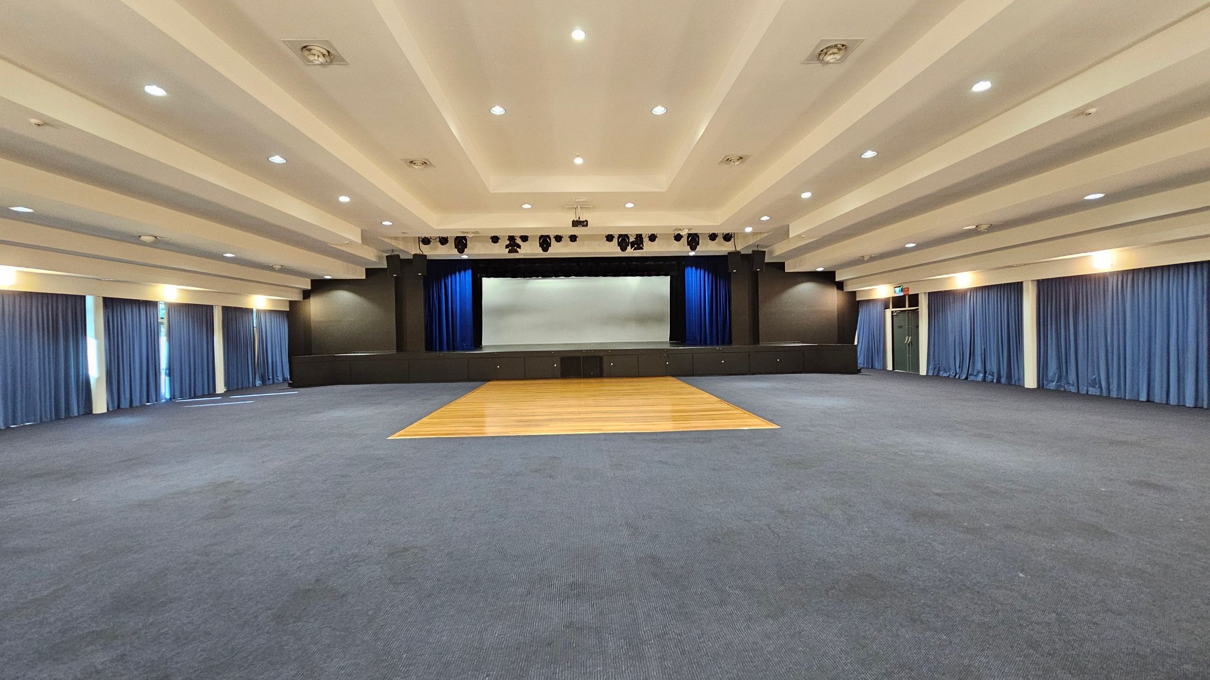 A spacious auditorium with a stage at the centre back, framed by blue curtains. The floor is mostly carpeted in dark grey, with a wooden section in front of the stage. The ceiling is white with recessed lighting and visible beams. Blue curtains line the walls on both sides, with a door visible on the right. The room is well-lit, creating a bright and open atmosphere. A spacious auditorium with a stage at the centre back, framed by blue curtains. The floor is mostly carpeted in dark grey, with a wooden section in front of the stage. The ceiling is white with recessed lighting and visible beams. Blue curtains line the walls on both sides, with a door visible on the right. The room is well-lit, creating a bright and open atmosphere.