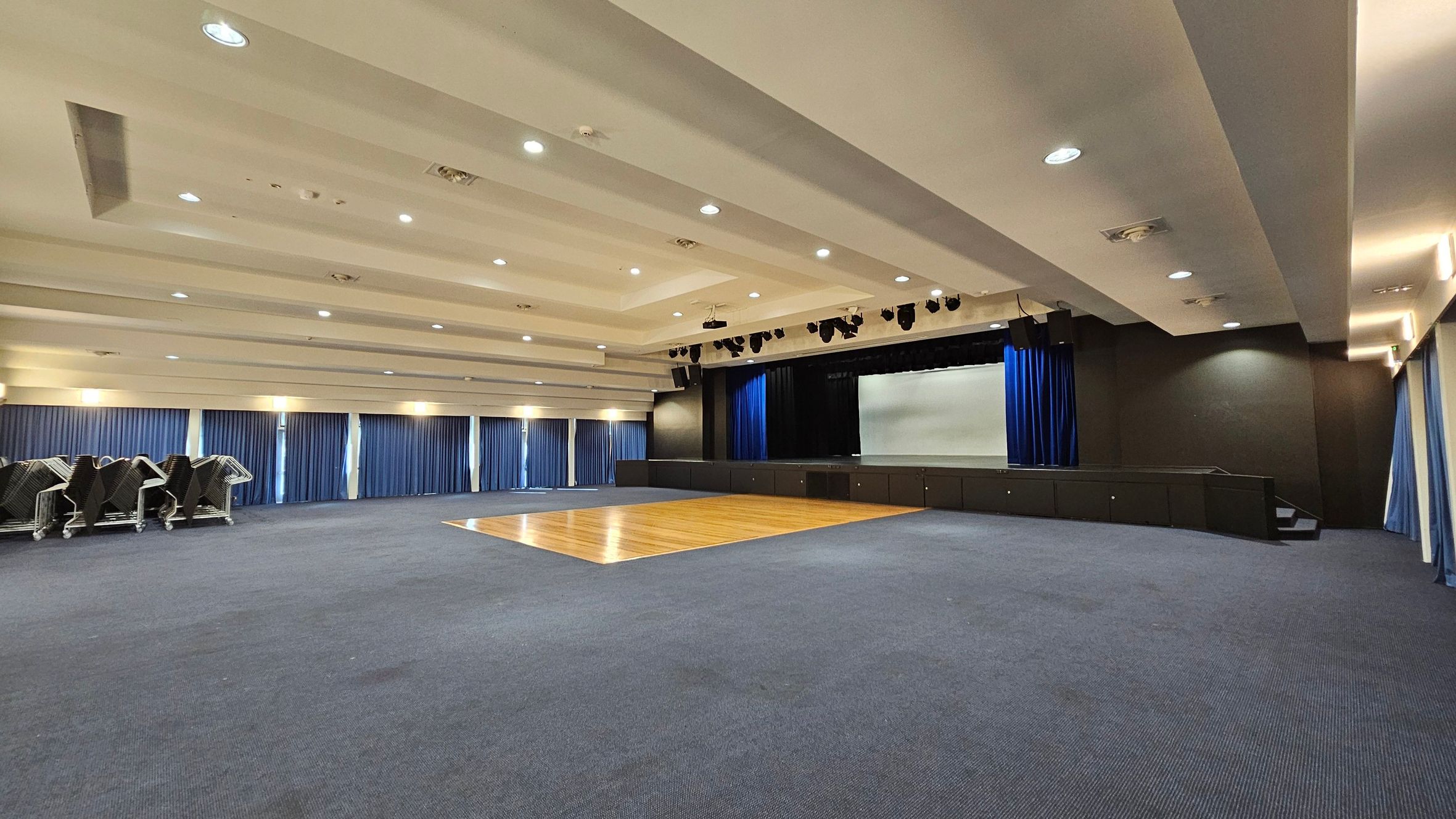 A spacious auditorium with a stage at the far end, featuring blue curtains and a large screen. The floor is mostly carpeted in blue, with a wooden section in front of the stage. On the left, there are stacks of chairs on trolleys. The ceiling is equipped with recessed lighting and speakers. The walls are lined with tall windows covered by blue curtains. A spacious auditorium with a stage at the far end, featuring blue curtains and a large screen. The floor is mostly carpeted in blue, with a wooden section in front of the stage. On the left, there are stacks of chairs on trolleys. The ceiling is equipped with recessed lighting and speakers. The walls are lined with tall windows covered by blue curtains.
