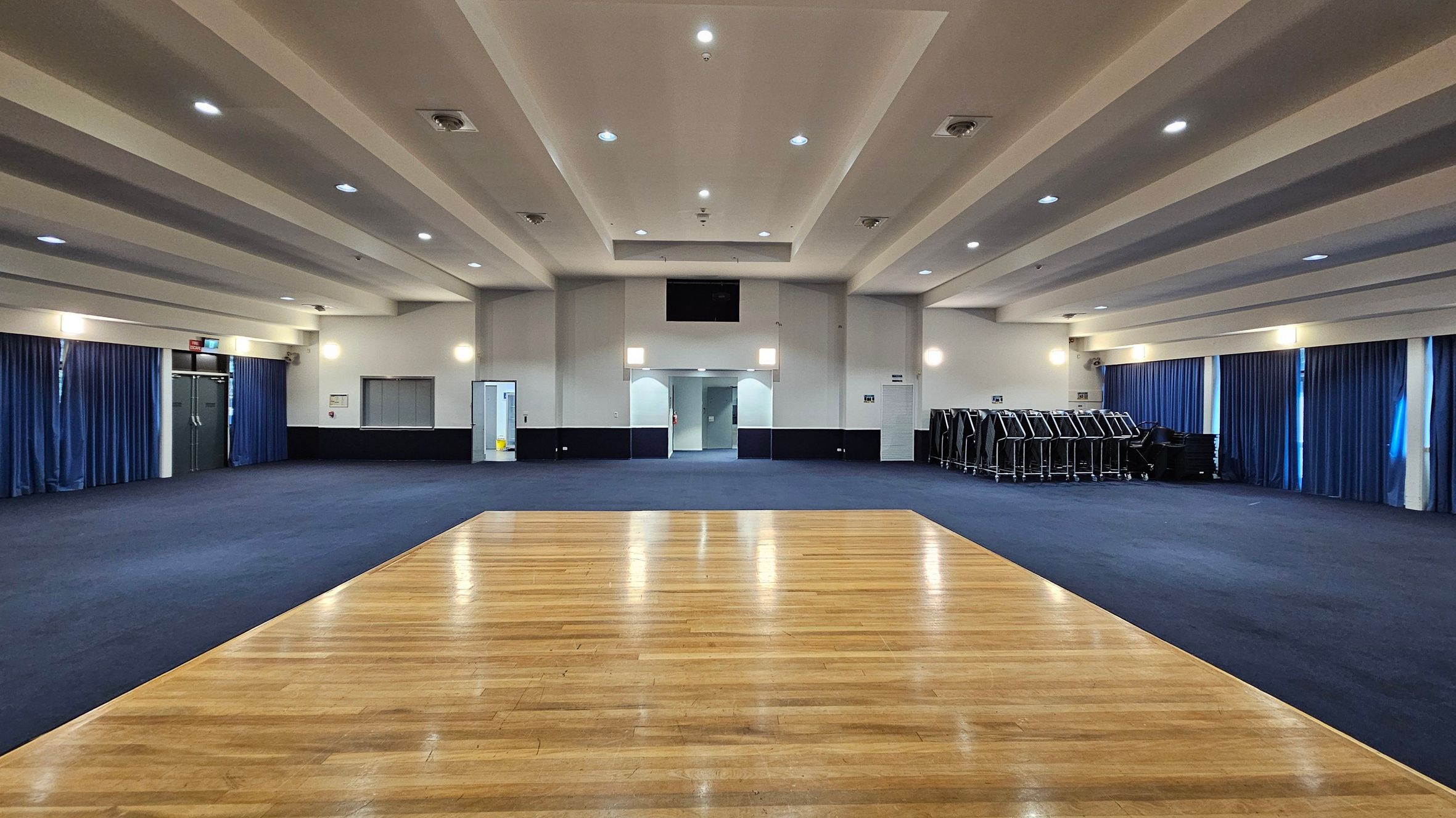 A large, empty room with a wooden floor section in the foreground and blue carpet covering the rest. The ceiling is white with recessed lighting. The walls are lined with blue curtains, and there are several doors and a stack of chairs on the right. The room is well-lit with wall-mounted lights and ceiling fixtures. A large, empty room with a wooden floor section in the foreground and blue carpet covering the rest. The ceiling is white with recessed lighting. The walls are lined with blue curtains, and there are several doors and a stack of chairs on the right. The room is well-lit with wall-mounted lights and ceiling fixtures.