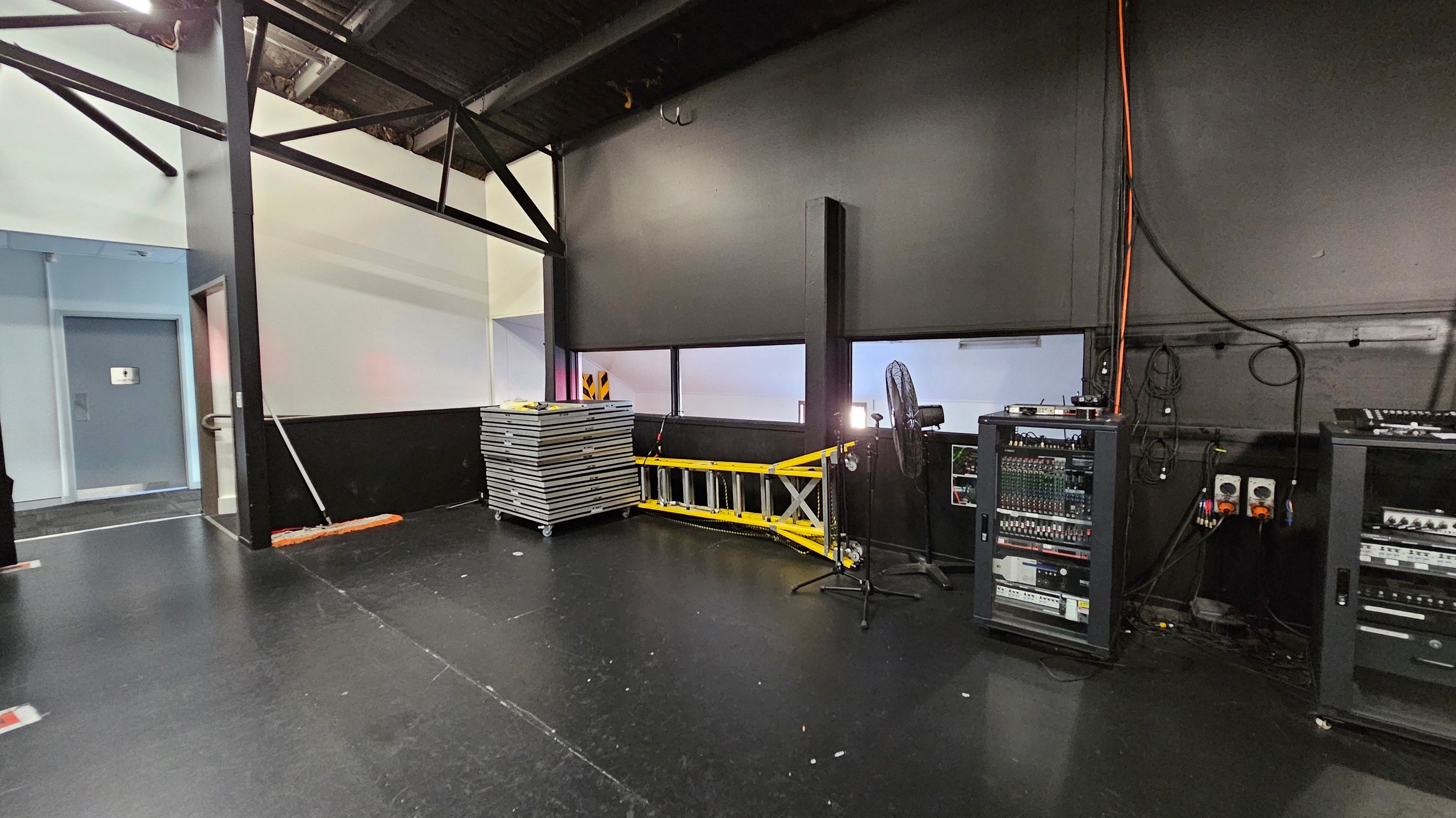 A backstage area with a black floor and ceiling. On the left, there's a blue door with a handle and a small sign. In the centre, a stack of metal platforms is next to a yellow ladder. A fan and microphone stand are nearby. On the right, there are two racks of audio equipment with various controls and cables. The walls are mostly black with some white sections, and orange cables run along the wall. A backstage area with a black floor and ceiling. On the left, there's a blue door with a handle and a small sign. In the centre, a stack of metal platforms is next to a yellow ladder. A fan and microphone stand are nearby. On the right, there are two racks of audio equipment with various controls and cables. The walls are mostly black with some white sections, and orange cables run along the wall.