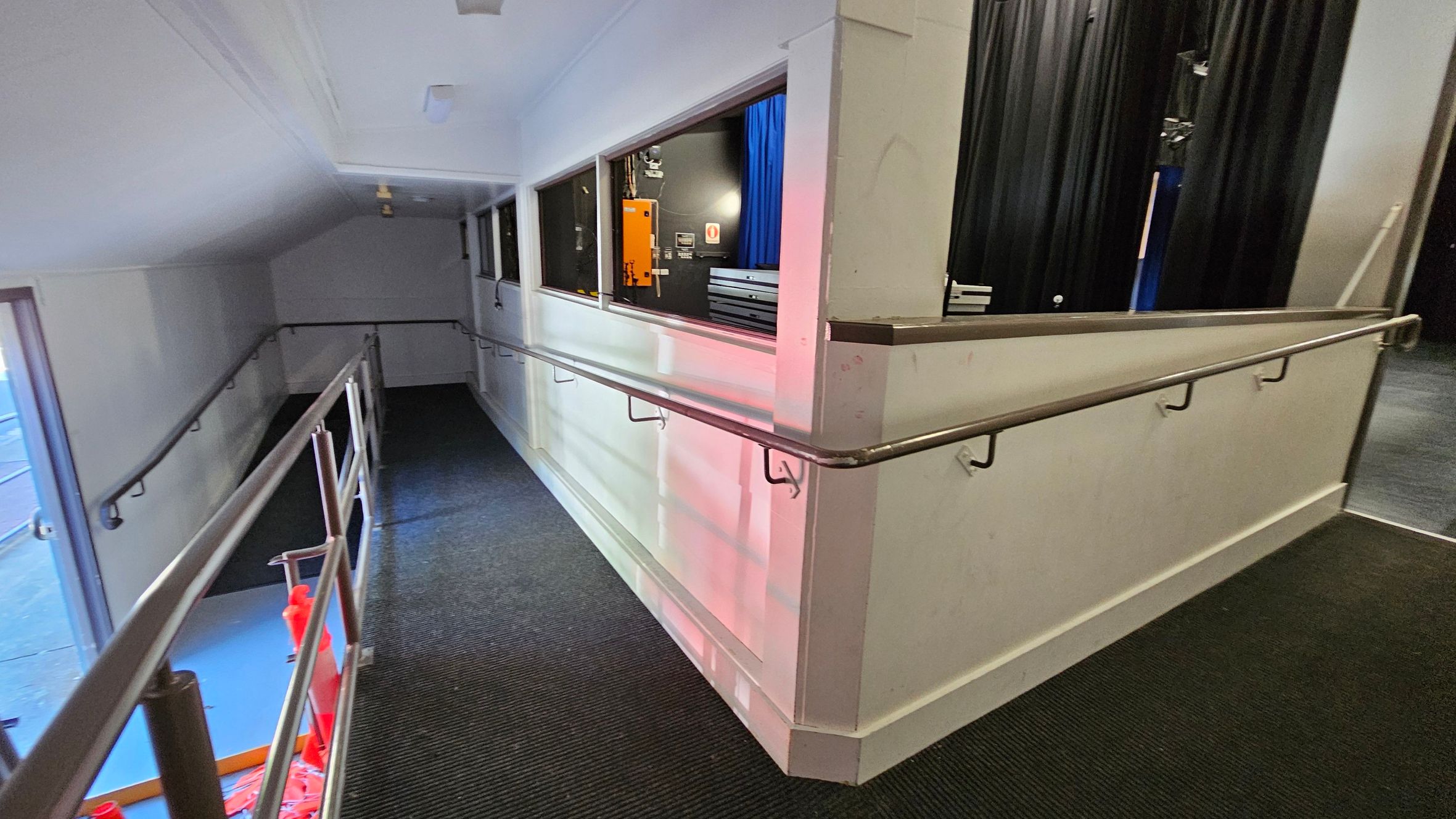A corridor with a sloped ramp and metal handrails on both sides. The left side has a window with a view of a blue and orange interior space. The right side features a wall with several windows, revealing a room with black curtains and some equipment. The floor is carpeted in dark grey, and the ceiling is white with a few light fixtures. A corridor with a sloped ramp and metal handrails on both sides. The left side has a window with a view of a blue and orange interior space. The right side features a wall with several windows, revealing a room with black curtains and some equipment. The floor is carpeted in dark grey, and the ceiling is white with a few light fixtures.