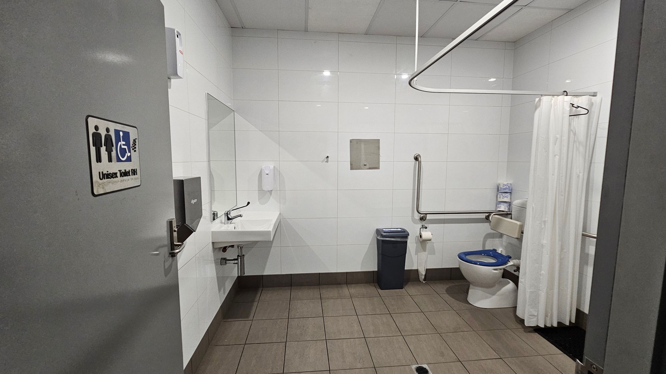 A public restroom with a grey door on the left, featuring a unisex and accessible toilet sign. Inside, the walls are covered with white tiles. On the left, a white sink with a soap dispenser and a paper towel dispenser is mounted on the wall. In the centre, a waste bin is placed on the brown-tiled floor. On the right, a toilet with a blue seat is equipped with grab bars and a white curtain. The ceiling is made of white panels. A public restroom with a grey door on the left, featuring a unisex and accessible toilet sign. Inside, the walls are covered with white tiles. On the left, a white sink with a soap dispenser and a paper towel dispenser is mounted on the wall. In the centre, a waste bin is placed on the brown-tiled floor. On the right, a toilet with a blue seat is equipped with grab bars and a white curtain. The ceiling is made of white panels.