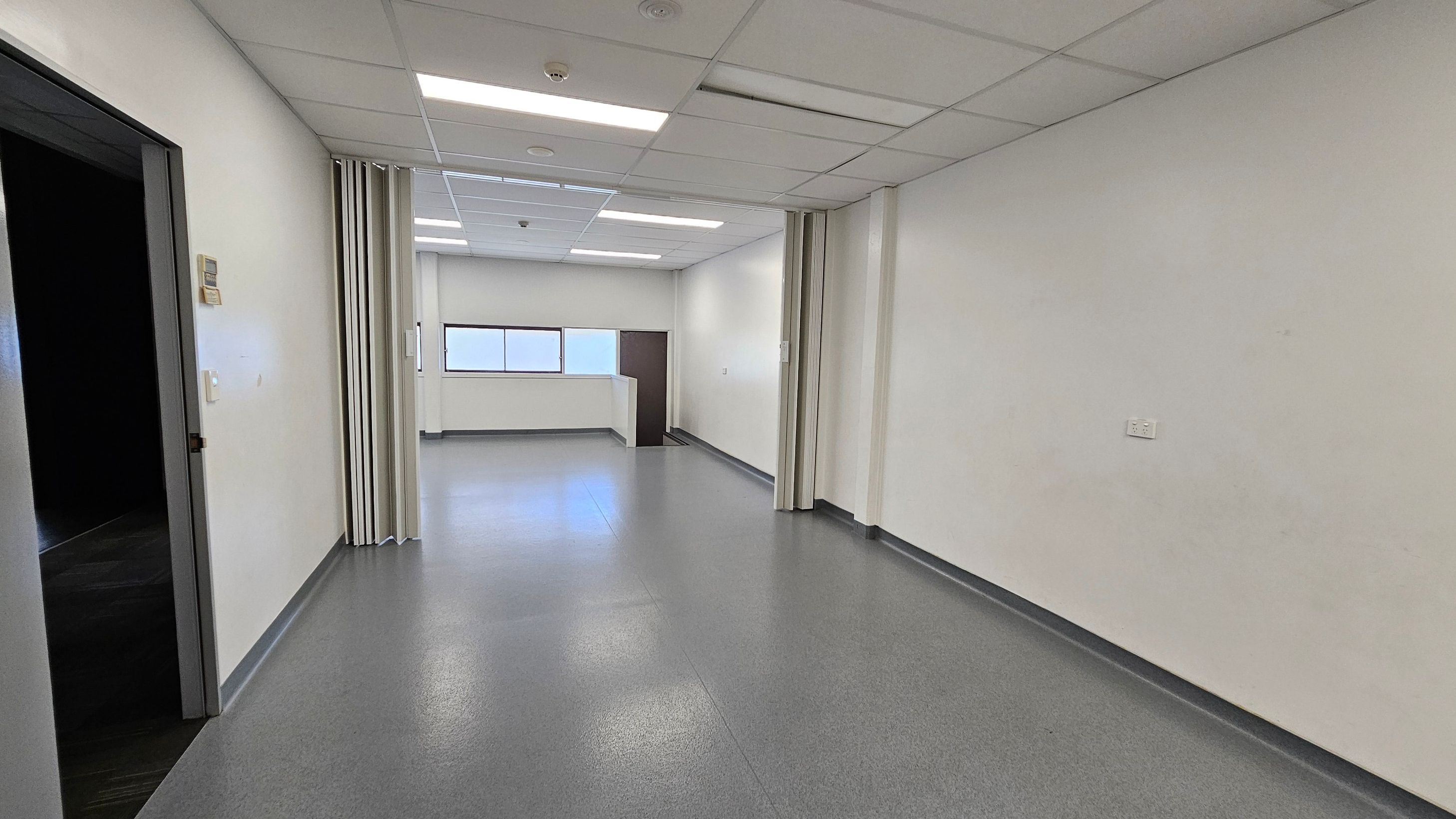 A corridor with grey flooring and white walls. On the left, there's a dark doorway and a wall-mounted control panel. In the centre, a partition with green and white panels partially divides the space. At the far end, a small window and a dark door are visible. The ceiling is fitted with white panels and fluorescent lights. A corridor with grey flooring and white walls. On the left, there's a dark doorway and a wall-mounted control panel. In the centre, a partition with green and white panels partially divides the space. At the far end, a small window and a dark door are visible. The ceiling is fitted with white panels and fluorescent lights.