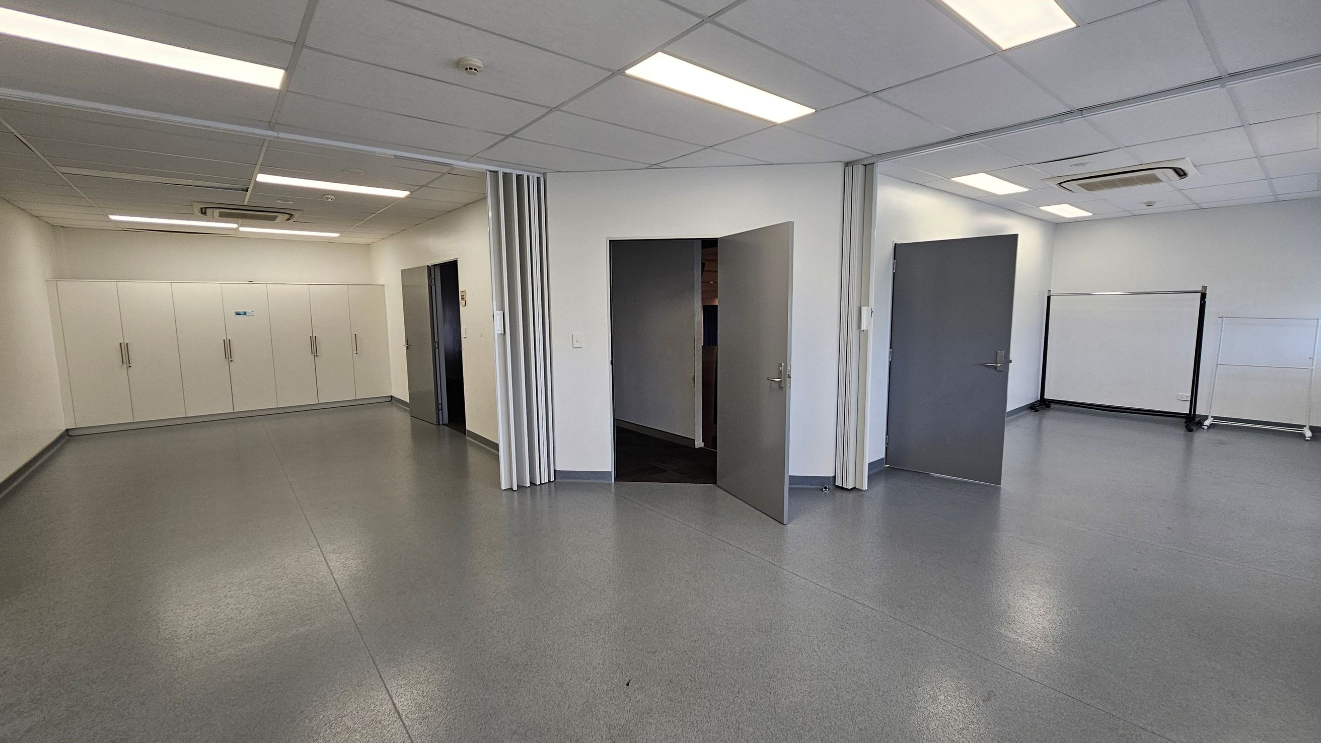 A room with a grey, speckled floor and a white ceiling with recessed lighting. On the left, there are white cabinets with handles along the wall. In the centre, two grey doors are open, leading to another area. On the right, a whiteboard on wheels is positioned against the wall. The ceiling features an air conditioning unit. The walls are plain and light-coloured, contributing to a clean and minimalistic appearance. A room with a grey, speckled floor and a white ceiling with recessed lighting. On the left, there are white cabinets with handles along the wall. In the centre, two grey doors are open, leading to another area. On the right, a whiteboard on wheels is positioned against the wall. The ceiling features an air conditioning unit. The walls are plain and light-coloured, contributing to a clean and minimalistic appearance.