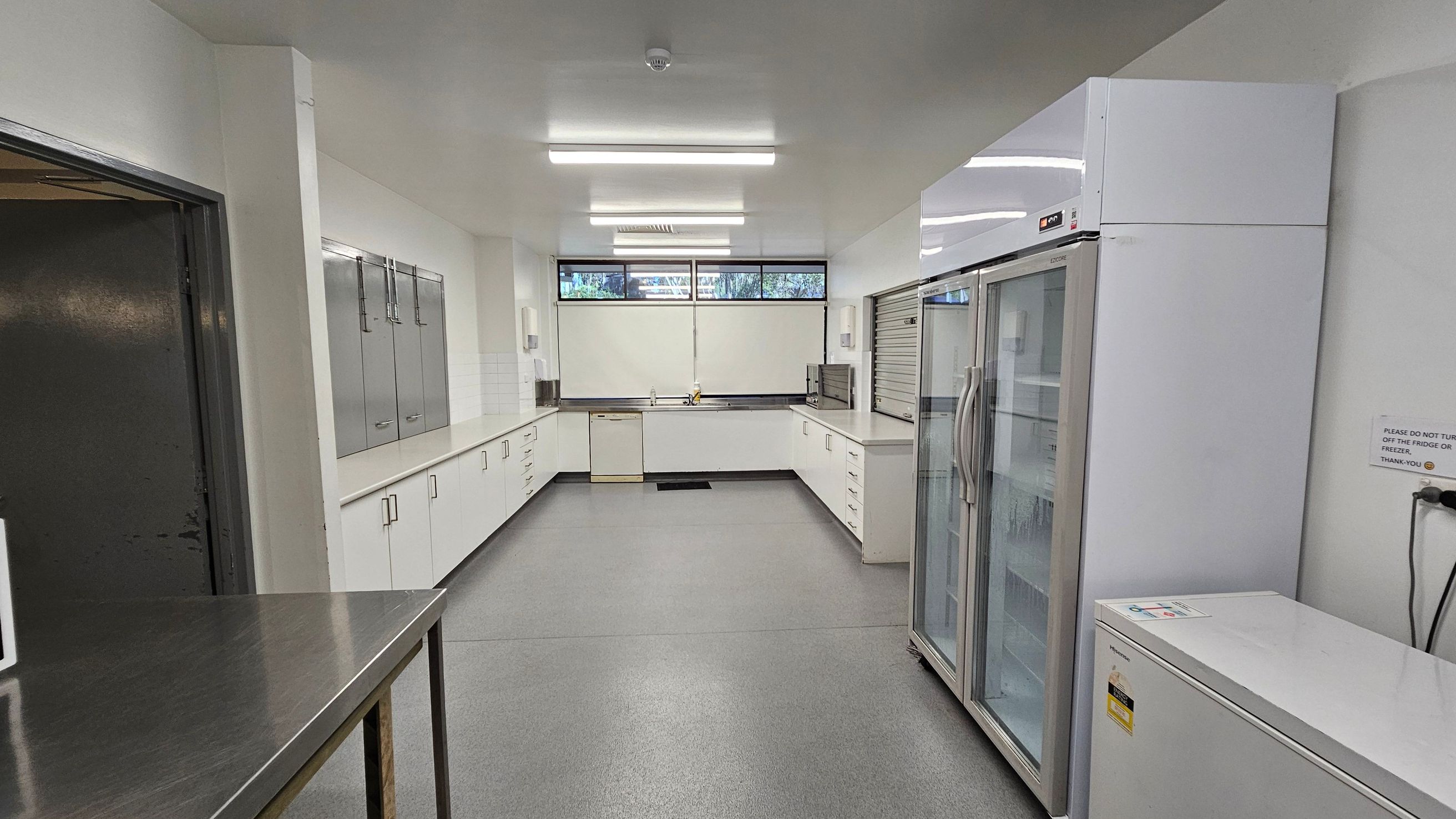 A clean, modern kitchen with a grey floor and white ceiling. On the left, there is a stainless steel table and a dark door. The right side features a large white refrigerator with glass doors and a chest freezer. The back wall has a long counter with white cabinets, a sink, and a dishwasher. Above the counter, there are windows with blinds partially drawn. Fluorescent lights are mounted on the ceiling. A clean, modern kitchen with a grey floor and white ceiling. On the left, there is a stainless steel table and a dark door. The right side features a large white refrigerator with glass doors and a chest freezer. The back wall has a long counter with white cabinets, a sink, and a dishwasher. Above the counter, there are windows with blinds partially drawn. Fluorescent lights are mounted on the ceiling.