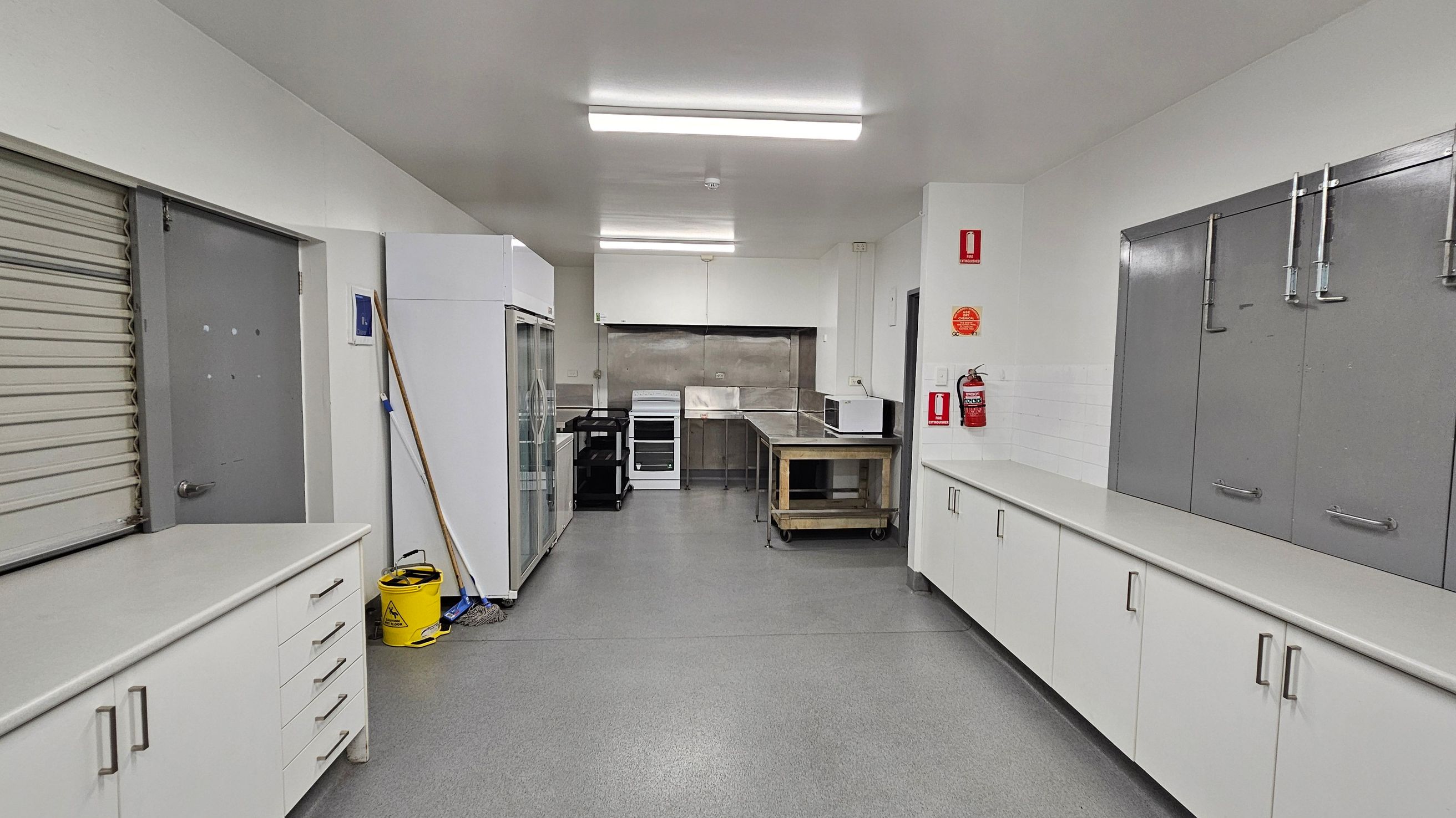 A commercial kitchen with a grey floor and white ceiling. On the left, there is a closed roller door, a grey door, and cleaning equipment including a mop and bucket. In the centre, there is a stainless steel counter, a stove, and a microwave on a trolley. On the right, there are white cabinets with grey doors above, and a fire extinguisher mounted on the wall. Fluorescent lights illuminate the space. A commercial kitchen with a grey floor and white ceiling. On the left, there is a closed roller door, a grey door, and cleaning equipment including a mop and bucket. In the centre, there is a stainless steel counter, a stove, and a microwave on a trolley. On the right, there are white cabinets with grey doors above, and a fire extinguisher mounted on the wall. Fluorescent lights illuminate the space.