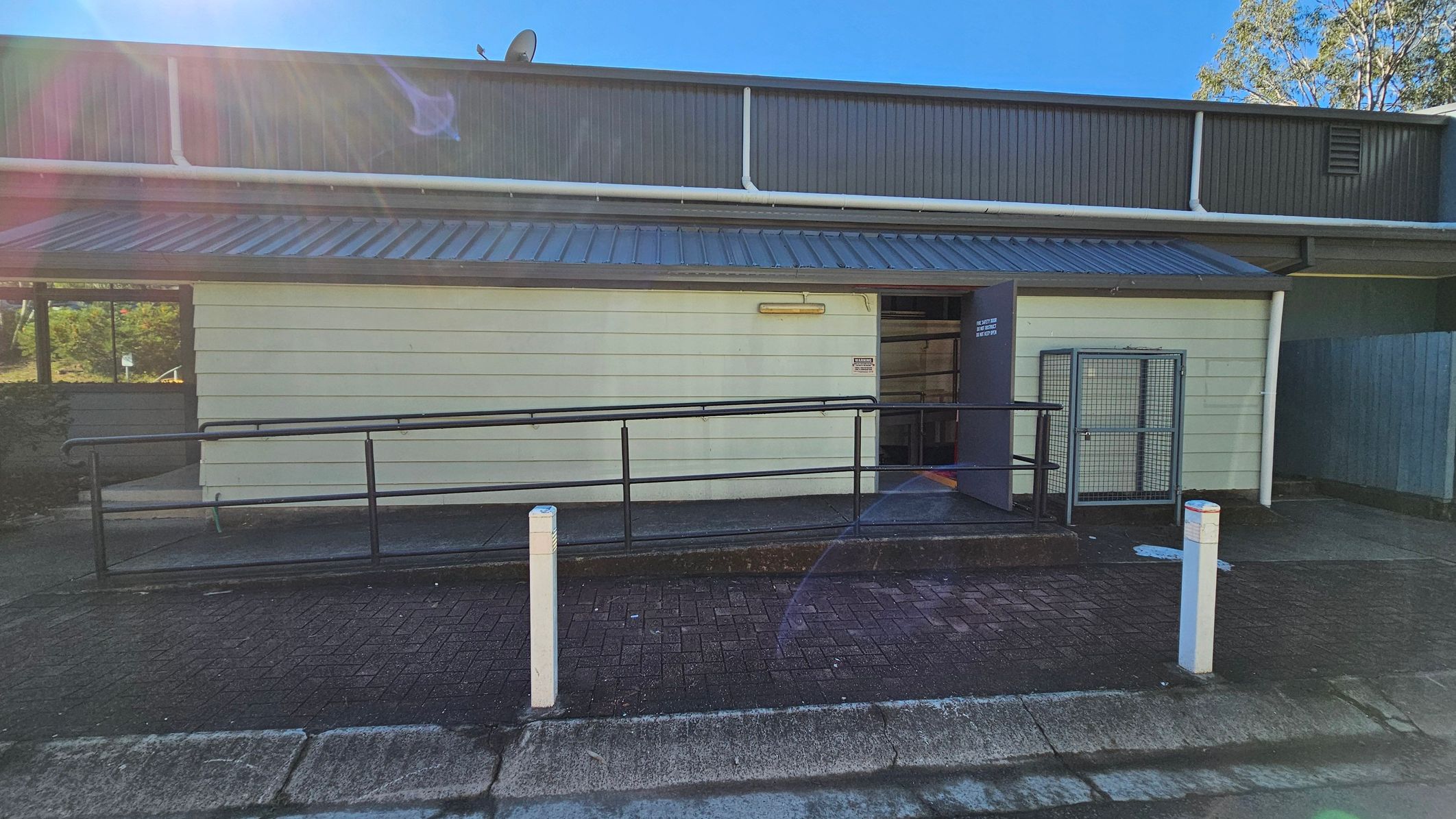 Single-storey building with a corrugated metal roof and light-coloured horizontal siding. A black metal railing runs along a ramp leading to an open door on the right. A small, cage-like structure is adjacent to the door. Two white bollards are positioned in front of the building on a paved surface. A window on the left reflects greenery. The upper part of the building has vertical cladding and a visible gutter system. Trees are partially visible in the background. Single-storey building with a corrugated metal roof and light-coloured horizontal siding. A black metal railing runs along a ramp leading to an open door on the right. A small, cage-like structure is adjacent to the door. Two white bollards are positioned in front of the building on a paved surface. A window on the left reflects greenery. The upper part of the building has vertical cladding and a visible gutter system. Trees are partially visible in the background.