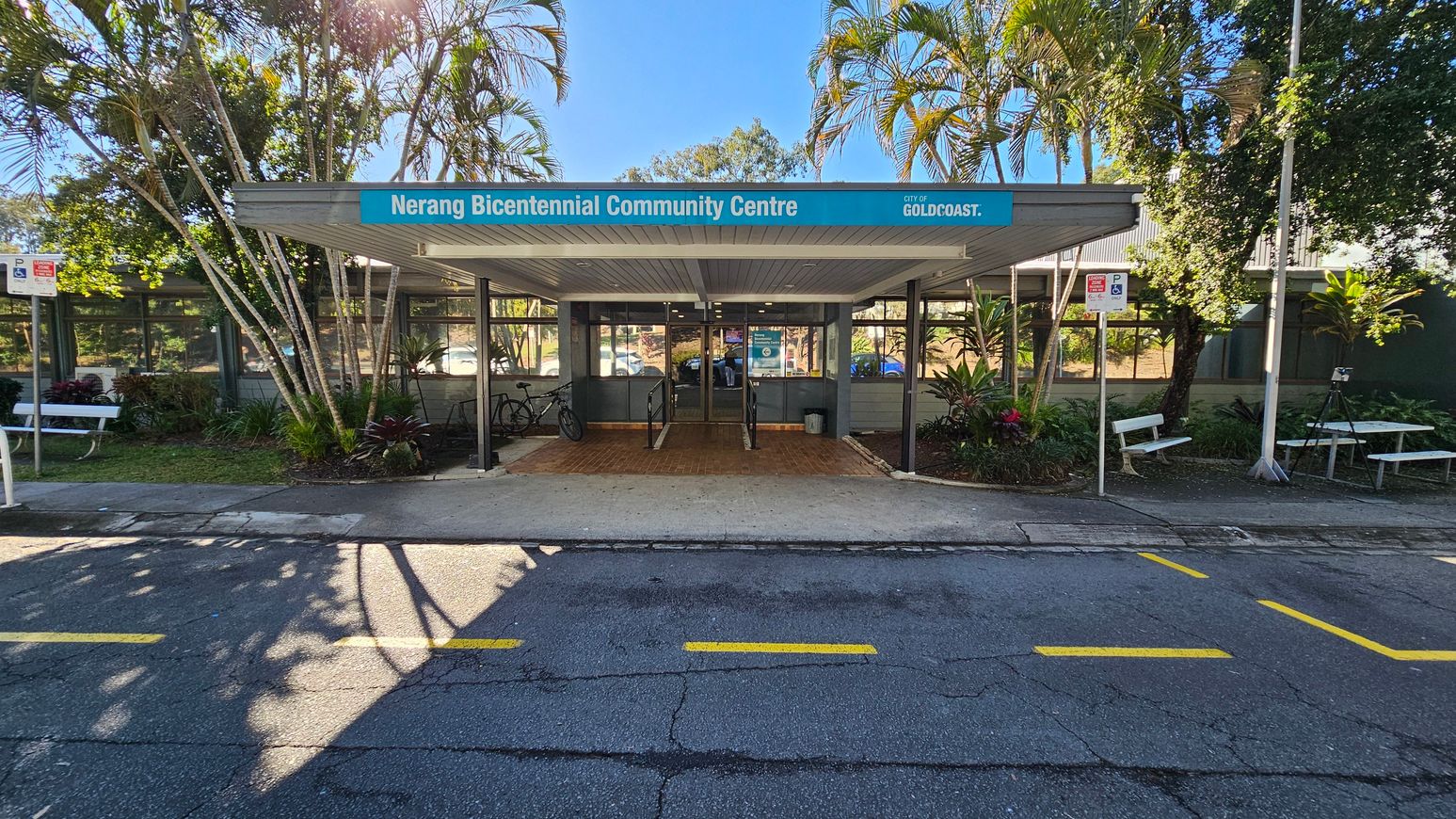 Entrance to the Nerang Bicentennial Community Centre with a blue sign above. The building has large windows and a covered walkway leading to the entrance. Bicycles are parked on the left. Palm trees and other greenery surround the area. Two white benches are on either side, with a picnic table on the right. The foreground shows a road with yellow markings and shadows from the trees. Entrance to the Nerang Bicentennial Community Centre with a blue sign above. The building has large windows and a covered walkway leading to the entrance. Bicycles are parked on the left. Palm trees and other greenery surround the area. Two white benches are on either side, with a picnic table on the right. The foreground shows a road with yellow markings and shadows from the trees.