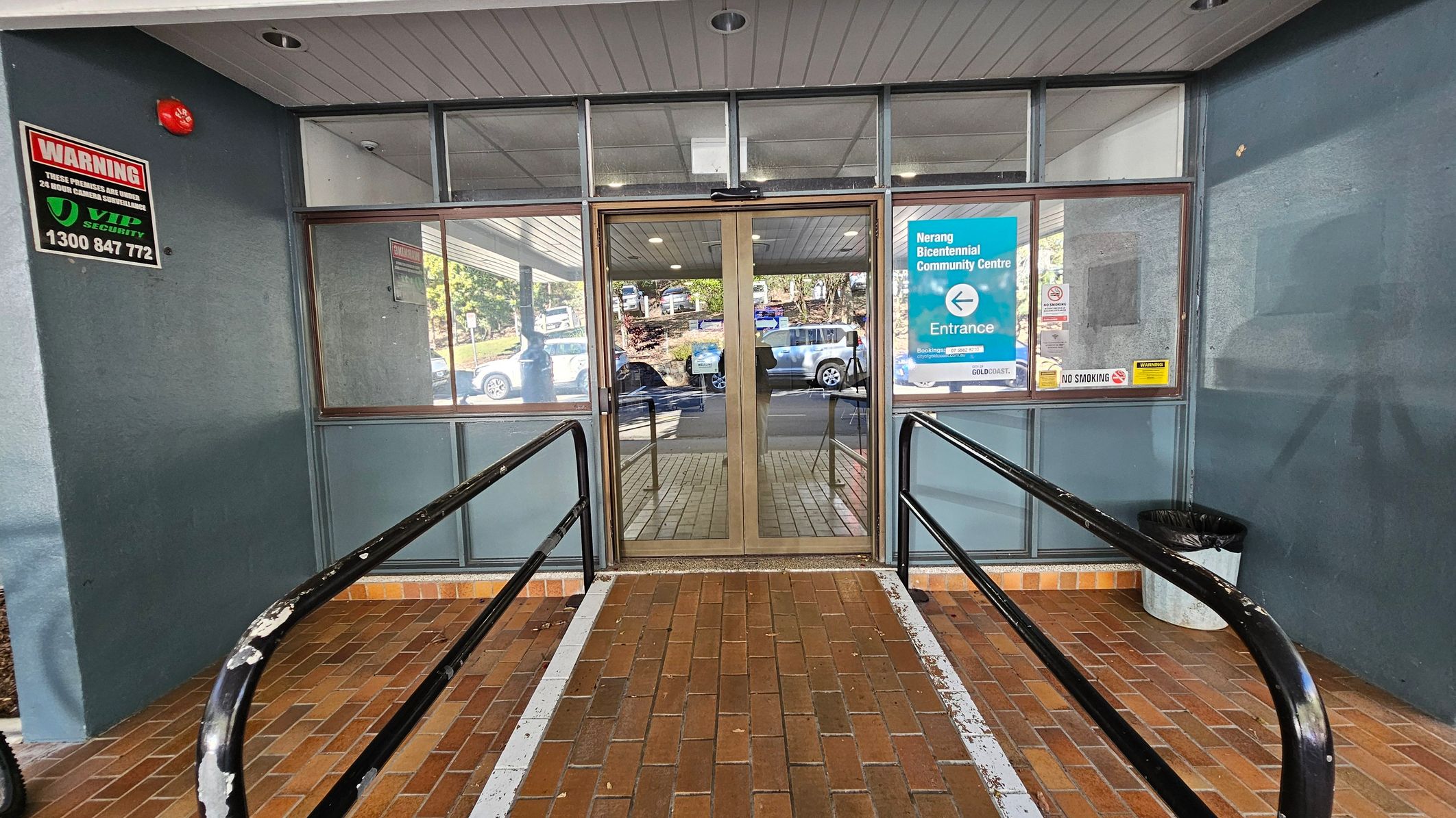 Glass entrance doors with metal frames are in the centre, flanked by large windows. A sign on the right reads "Nerang Bicentennial Community Centre" with an arrow indicating the entrance. A "No Smoking" sign is also visible. On the left, a security warning sign is displayed. The floor is made of brown bricks, and there are black handrails on either side of a ramp leading to the doors. A grey ceiling with recessed lights is above, and a black bin is on the right. Glass entrance doors with metal frames are in the centre, flanked by large windows. A sign on the right reads "Nerang Bicentennial Community Centre" with an arrow indicating the entrance. A "No Smoking" sign is also visible. On the left, a security warning sign is displayed. The floor is made of brown bricks, and there are black handrails on either side of a ramp leading to the doors. A grey ceiling with recessed lights is above, and a black bin is on the right.