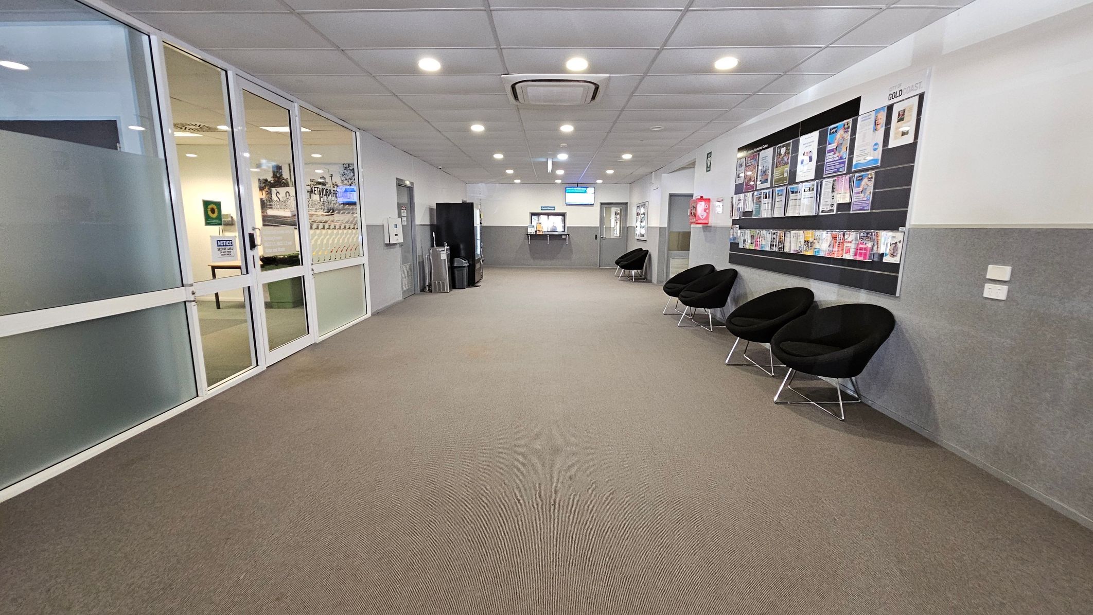 A hallway with a brown carpet and a white tiled ceiling featuring recessed lights. On the left, there are glass doors with a view into an adjacent room. On the right, a wall-mounted bulletin board displays various pamphlets and notices above a row of black chairs. In the background, a counter with a digital screen is visible, flanked by more chairs and a vending machine. A hallway with a brown carpet and a white tiled ceiling featuring recessed lights. On the left, there are glass doors with a view into an adjacent room. On the right, a wall-mounted bulletin board displays various pamphlets and notices above a row of black chairs. In the background, a counter with a digital screen is visible, flanked by more chairs and a vending machine.