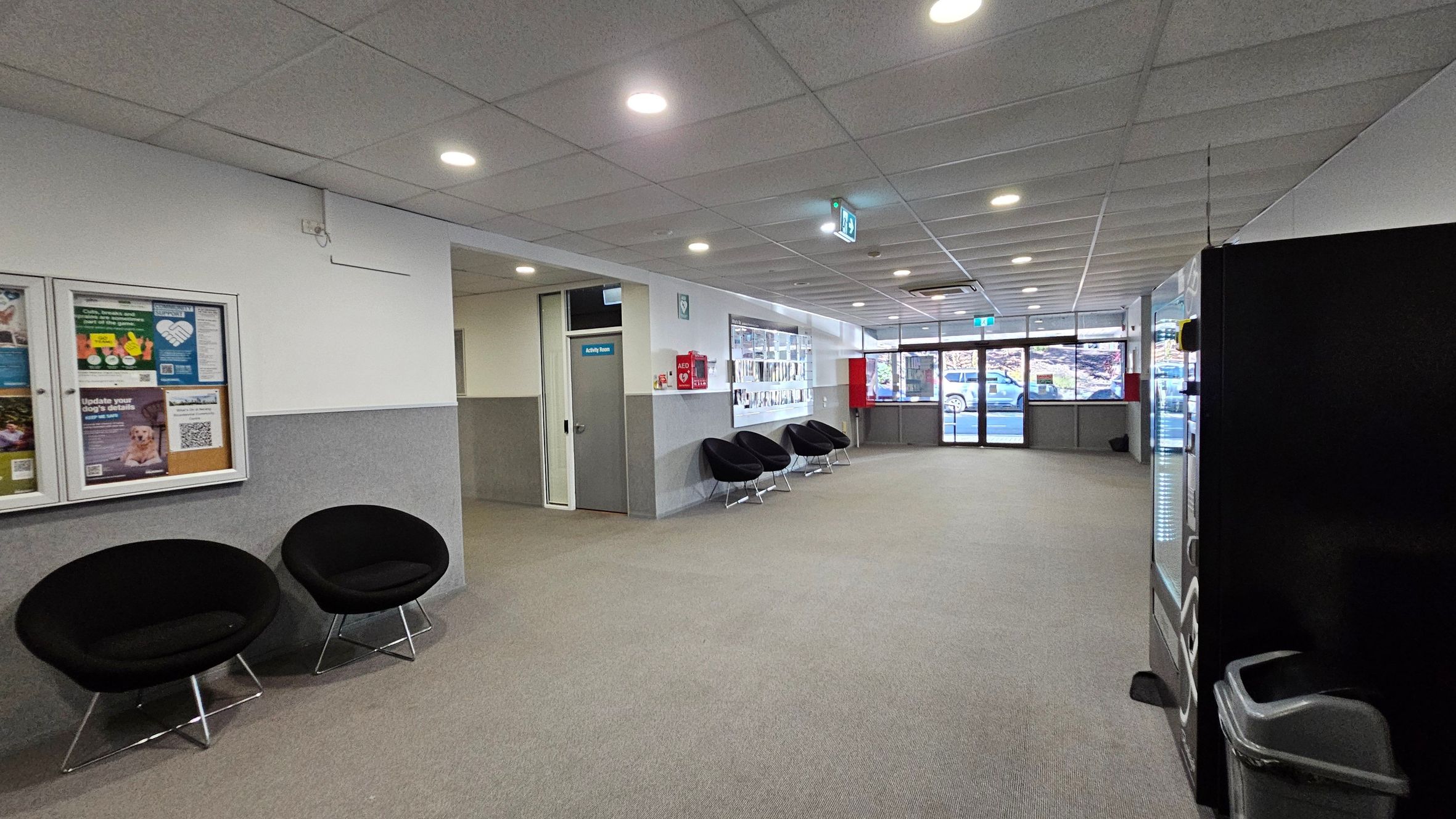 A spacious waiting area with grey carpet and a tiled ceiling with recessed lighting. On the left, there are two black circular chairs and a bulletin board with various notices. In the middle, more black chairs are lined up against the wall. A door labelled "Activity Room" is visible. On the right, a vending machine and a waste bin are present. The background features large windows and glass doors, showing a view of parked cars outside. A spacious waiting area with grey carpet and a tiled ceiling with recessed lighting. On the left, there are two black circular chairs and a bulletin board with various notices. In the middle, more black chairs are lined up against the wall. A door labelled "Activity Room" is visible. On the right, a vending machine and a waste bin are present. The background features large windows and glass doors, showing a view of parked cars outside.