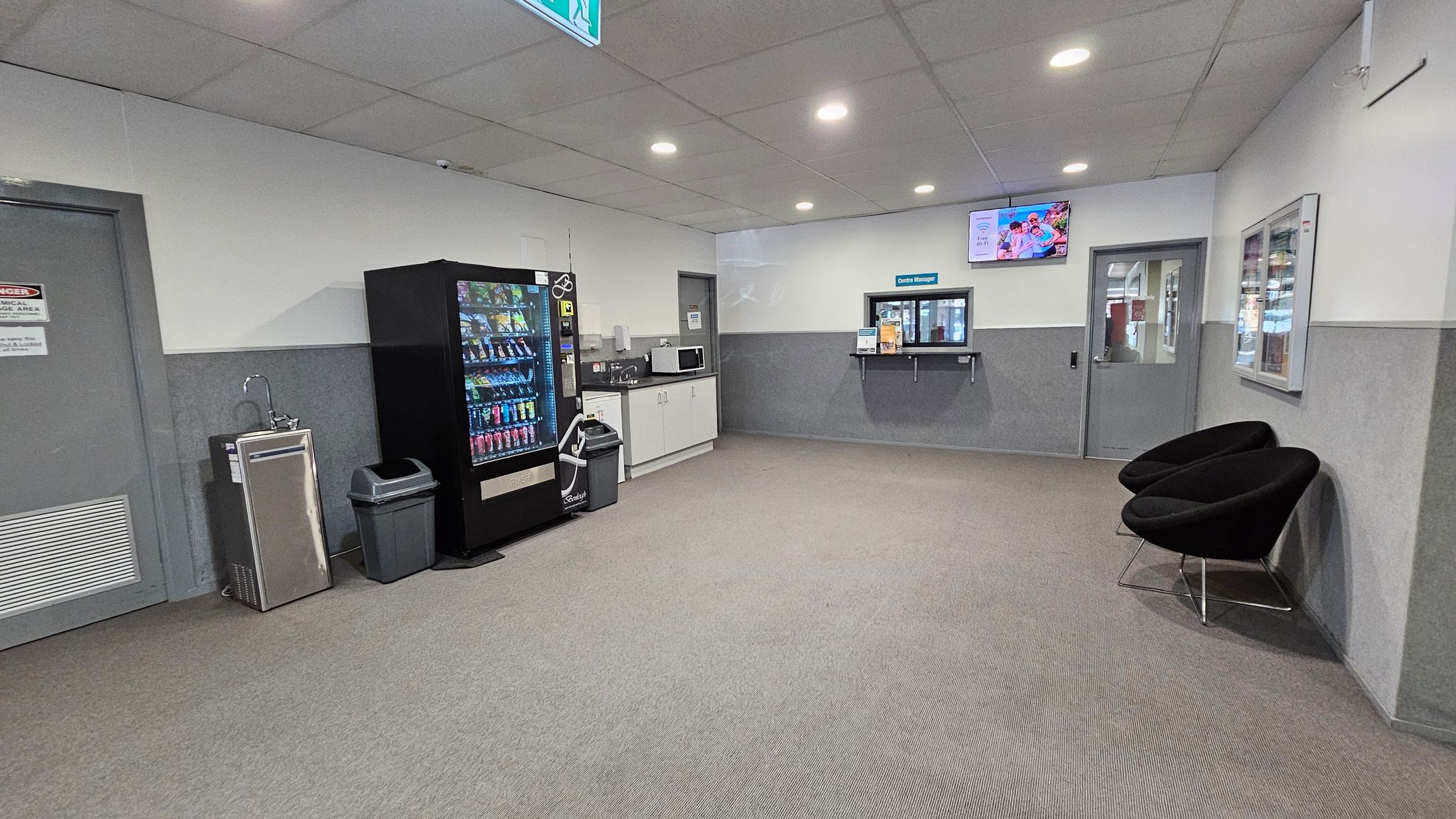 A waiting area with a grey carpeted floor and a tiled ceiling with recessed lights. On the left, there's a water fountain, a vending machine, and a waste bin. A countertop with a microwave and cabinets is nearby. In the centre, a window labelled "Centre Manager" is set into the wall. On the right, two black chairs are positioned against the wall. A television is mounted above the manager's window, and a door is visible on the far right. A waiting area with a grey carpeted floor and a tiled ceiling with recessed lights. On the left, there's a water fountain, a vending machine, and a waste bin. A countertop with a microwave and cabinets is nearby. In the centre, a window labelled "Centre Manager" is set into the wall. On the right, two black chairs are positioned against the wall. A television is mounted above the manager's window, and a door is visible on the far right.