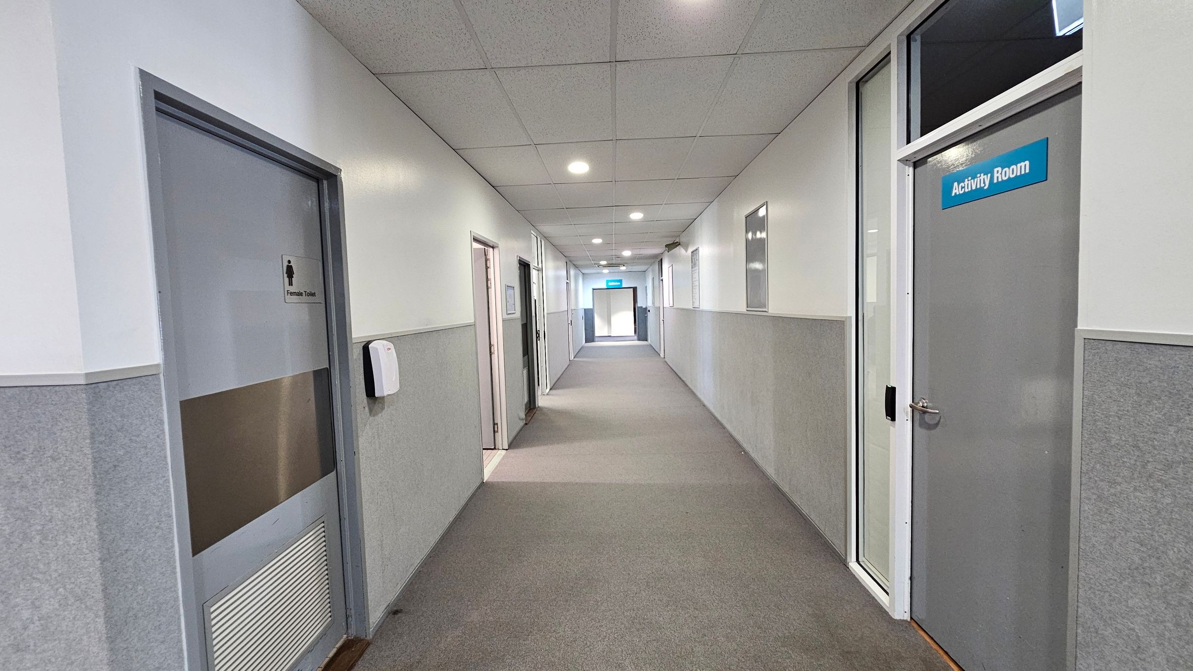A corridor with grey carpeted flooring and a tiled ceiling with recessed lights. On the left, there is a door labelled "Female Toilet" and a wall-mounted dispenser. On the right, a door marked "Activity Room" is visible. The walls are half-panelled with grey and white. At the end of the corridor, a blue sign is visible above a door, indicating an exit or another room. A corridor with grey carpeted flooring and a tiled ceiling with recessed lights. On the left, there is a door labelled "Female Toilet" and a wall-mounted dispenser. On the right, a door marked "Activity Room" is visible. The walls are half-panelled with grey and white. At the end of the corridor, a blue sign is visible above a door, indicating an exit or another room.