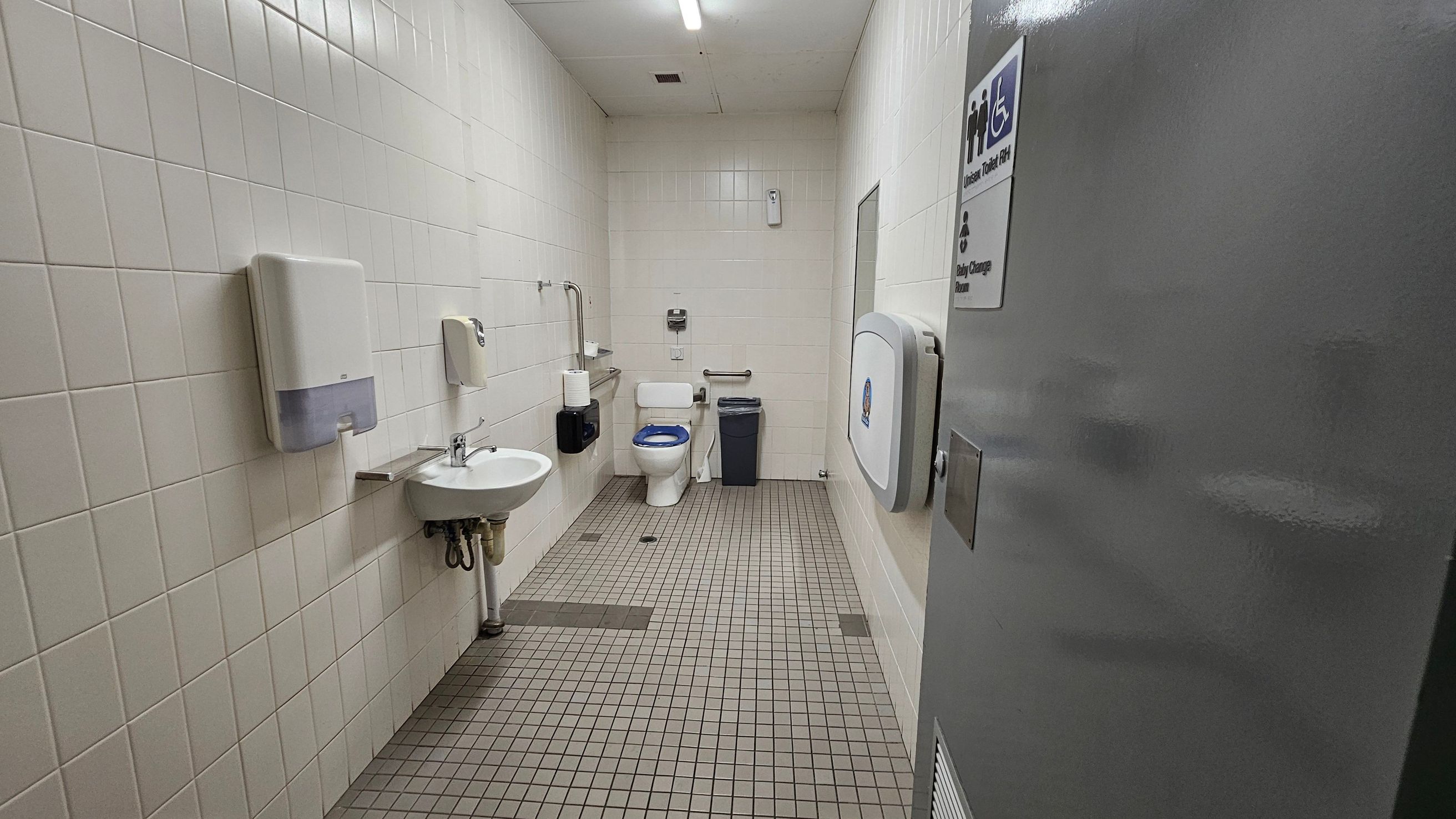 Accessible bathroom with white tiled walls and a green tiled floor. On the left, a sink with a soap dispenser and paper towel holder. In the centre, a toilet with grab bars and a bin nearby. On the right, a baby changing station is mounted on the wall. The ceiling is white with a fluorescent light. A sign on the door indicates it is a unisex and baby change facility. Accessible bathroom with white tiled walls and a green tiled floor. On the left, a sink with a soap dispenser and paper towel holder. In the centre, a toilet with grab bars and a bin nearby. On the right, a baby changing station is mounted on the wall. The ceiling is white with a fluorescent light. A sign on the door indicates it is a unisex and baby change facility.