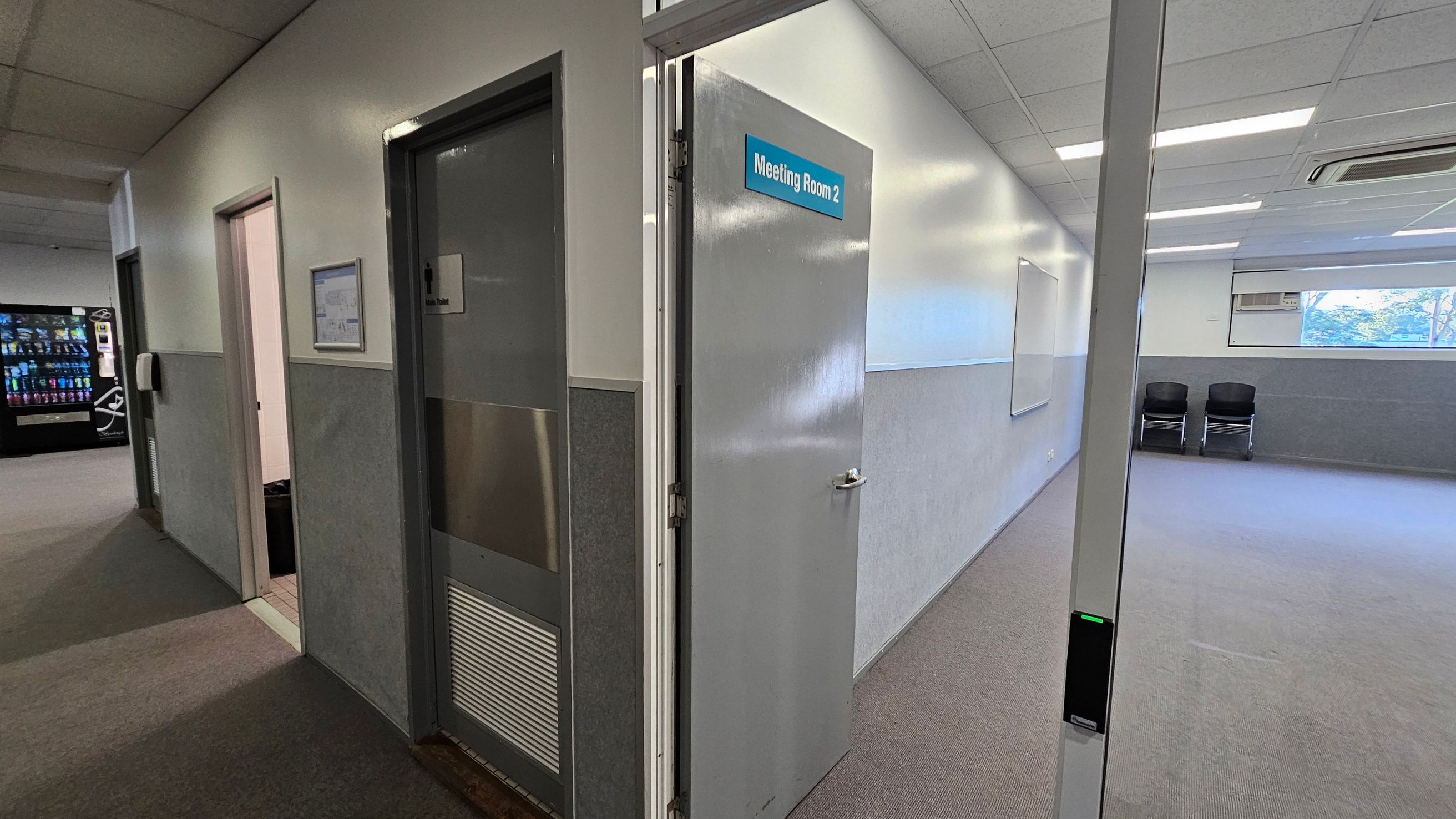 A hallway with grey carpet and a vending machine on the left. There are two doors on the left wall, one labelled "Meeting Room 2" and the other a bathroom. The meeting room door is open, revealing a room with a whiteboard, two black chairs, and a window. The walls are white with grey panelling, and the ceiling has fluorescent lights and an air conditioning unit. A hallway with grey carpet and a vending machine on the left. There are two doors on the left wall, one labelled "Meeting Room 2" and the other a bathroom. The meeting room door is open, revealing a room with a whiteboard, two black chairs, and a window. The walls are white with grey panelling, and the ceiling has fluorescent lights and an air conditioning unit.