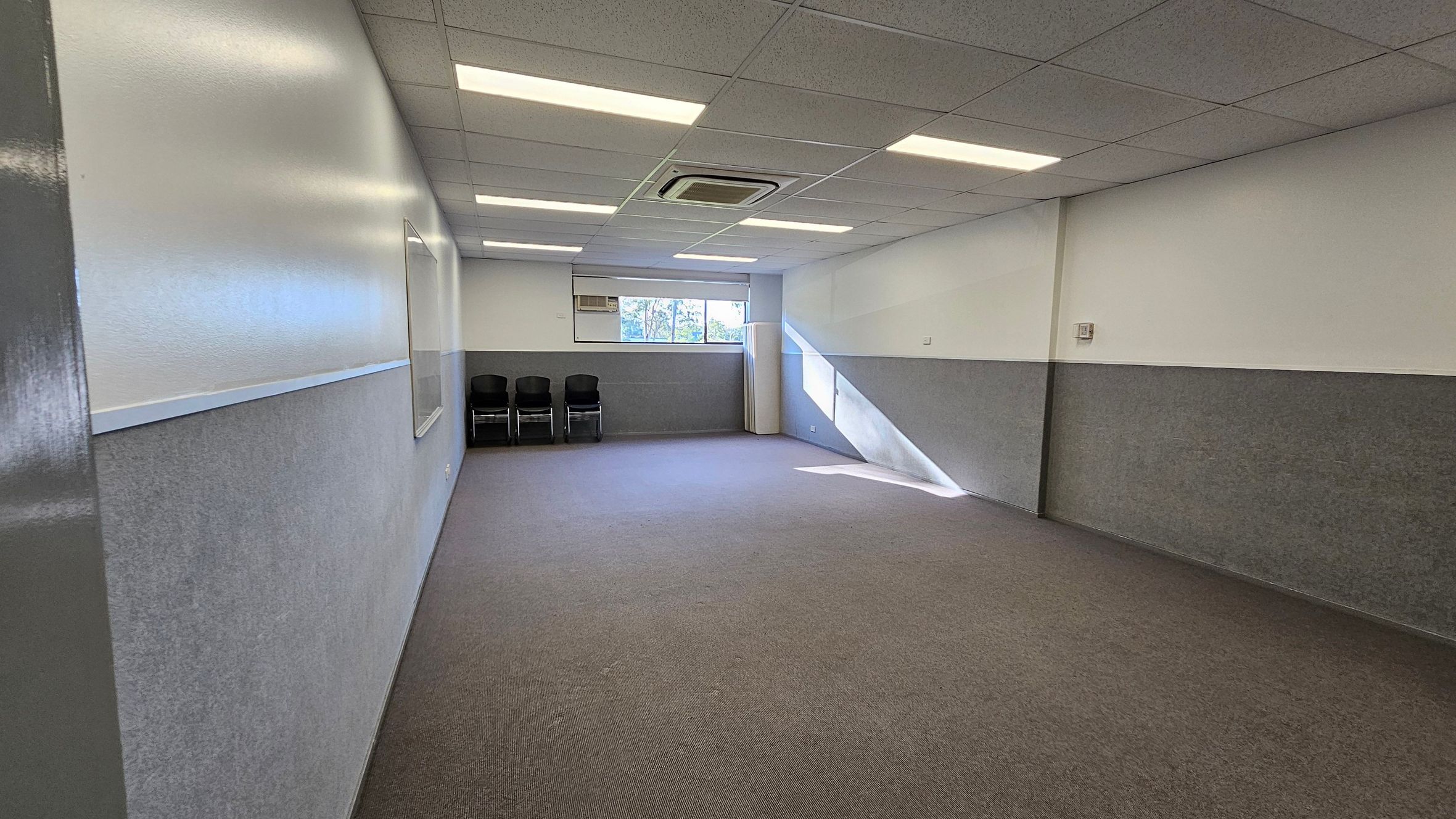 A rectangular room with grey carpeted flooring and a white ceiling featuring recessed lighting and an air conditioning unit. The walls are half-painted in grey and white. On the far wall, there is a window with a view of trees outside, allowing natural light to enter. Below the window, three black chairs are stacked. A whiteboard is mounted on the left wall. A rectangular room with grey carpeted flooring and a white ceiling featuring recessed lighting and an air conditioning unit. The walls are half-painted in grey and white. On the far wall, there is a window with a view of trees outside, allowing natural light to enter. Below the window, three black chairs are stacked. A whiteboard is mounted on the left wall.